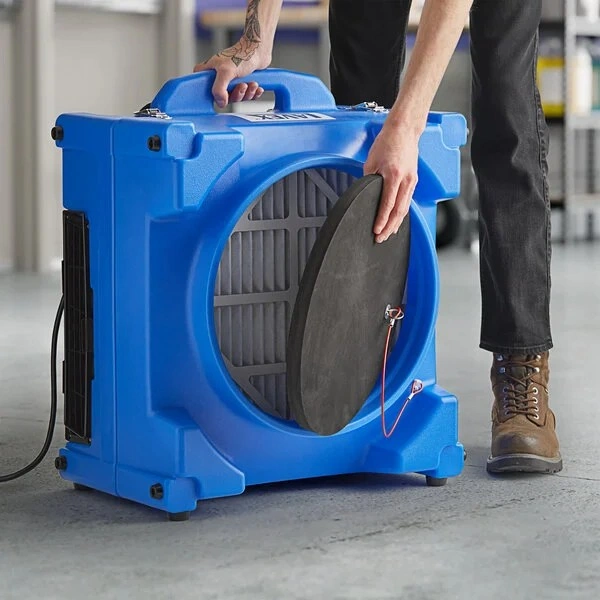 A person wearing black jeans and brown boots is holding the front filter cover of a large, blue, portable air scrubber unit.