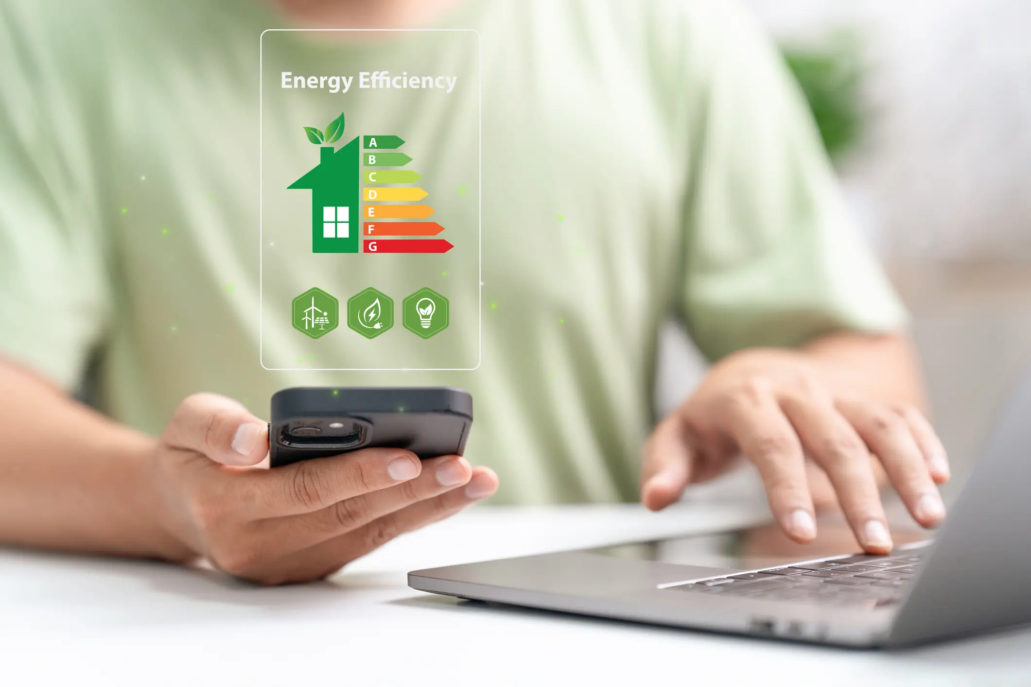 A person uses a smartphone to view a digital overlay showing a house energy efficiency rating chart and icons for green energy, with a laptop in the background.
