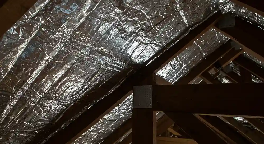 A close-up shot of a radiant barrier installed on the rafters of an attic. The silver, reflective surface is crinkled and stapled into place.