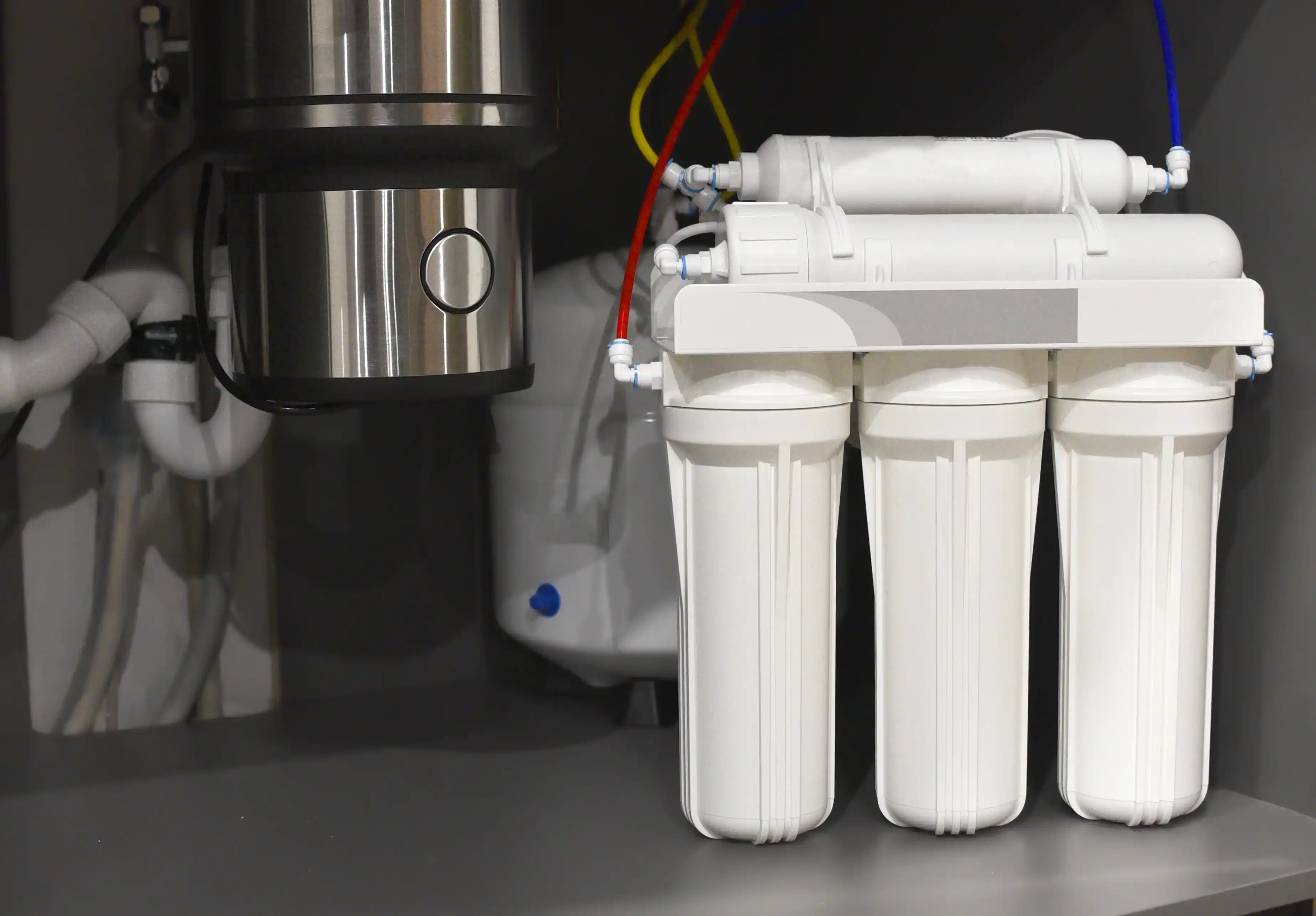 A three-stage reverse osmosis water filtration system is installed under a sink, featuring three white cylindrical filters and colorful red, yellow, and blue tubes.