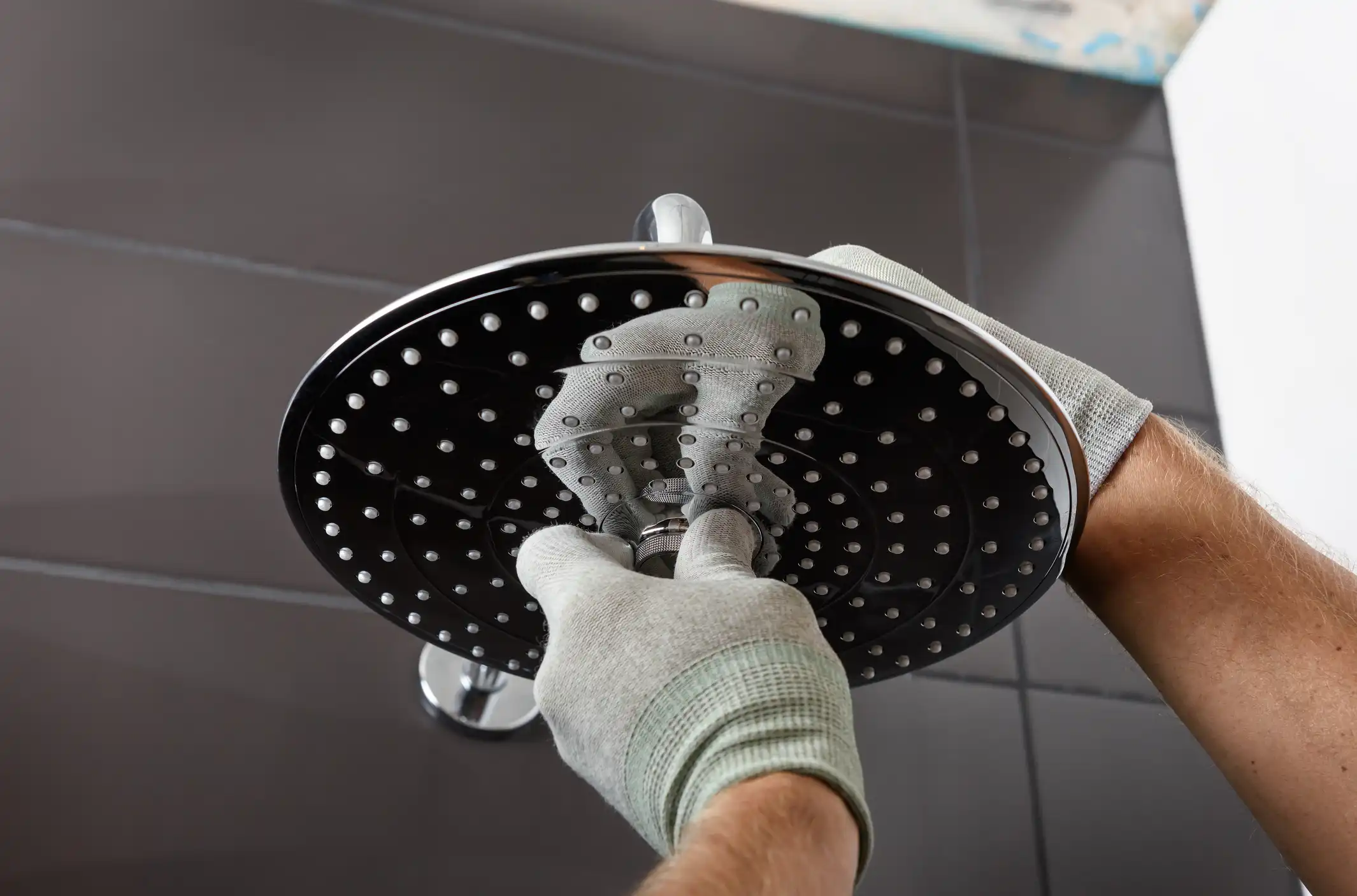 A person wearing grey work gloves is installing a large, circular, chrome showerhead.