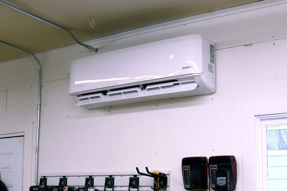 A white mini-split unit is mounted on a beige wall with a pipe wrapped in blue tape entering the wall