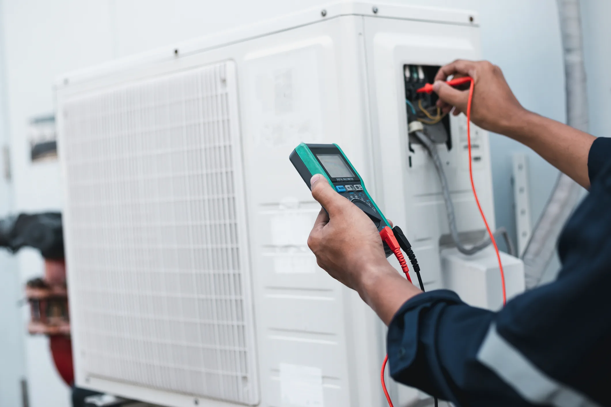 A close-up image shows a technician's hands performing an electrical diagnostic on a white outdoor air conditioning unit. The technician is wearing a dark uniform and holding a digital multimeter with a green screen. They are using the red test lead to check the wiring inside a small compartment of the AC unit, which has its cover removed to expose the electrical connections. The large, slotted fan grille of the condenser unit takes up the left side of the frame.