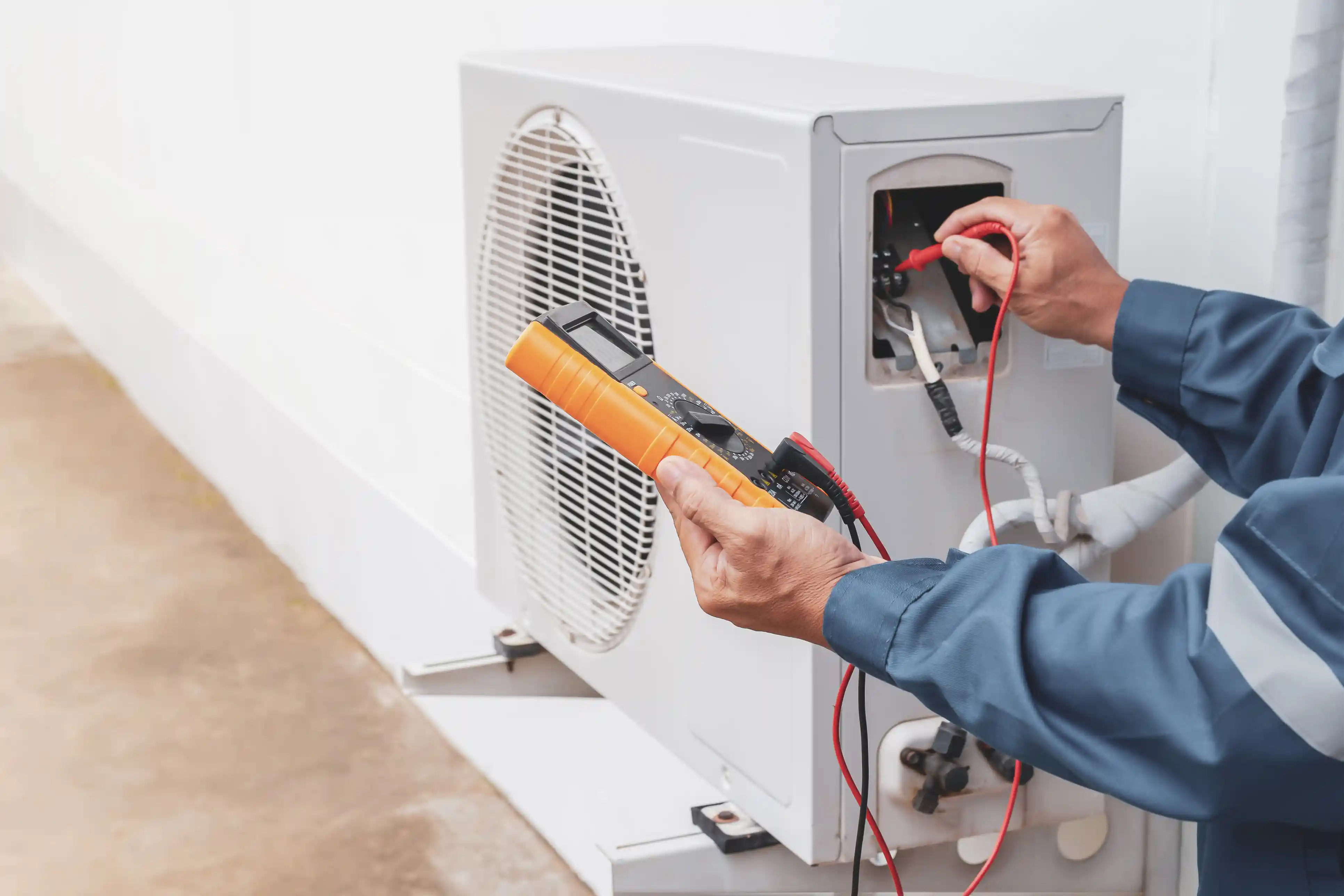 A close-up image shows a HVAC technician wearing a blue uniform using a digital multimeter with an orange casing to perform diagnostics on a white outdoor air conditioning unit. The technician's hands are holding the meter and inserting the red and black test leads into the unit's exposed electrical compartment. The AC unit, featuring a large circular fan grille, is mounted against a white wall, suggesting a professional electrical check during maintenance or repair.