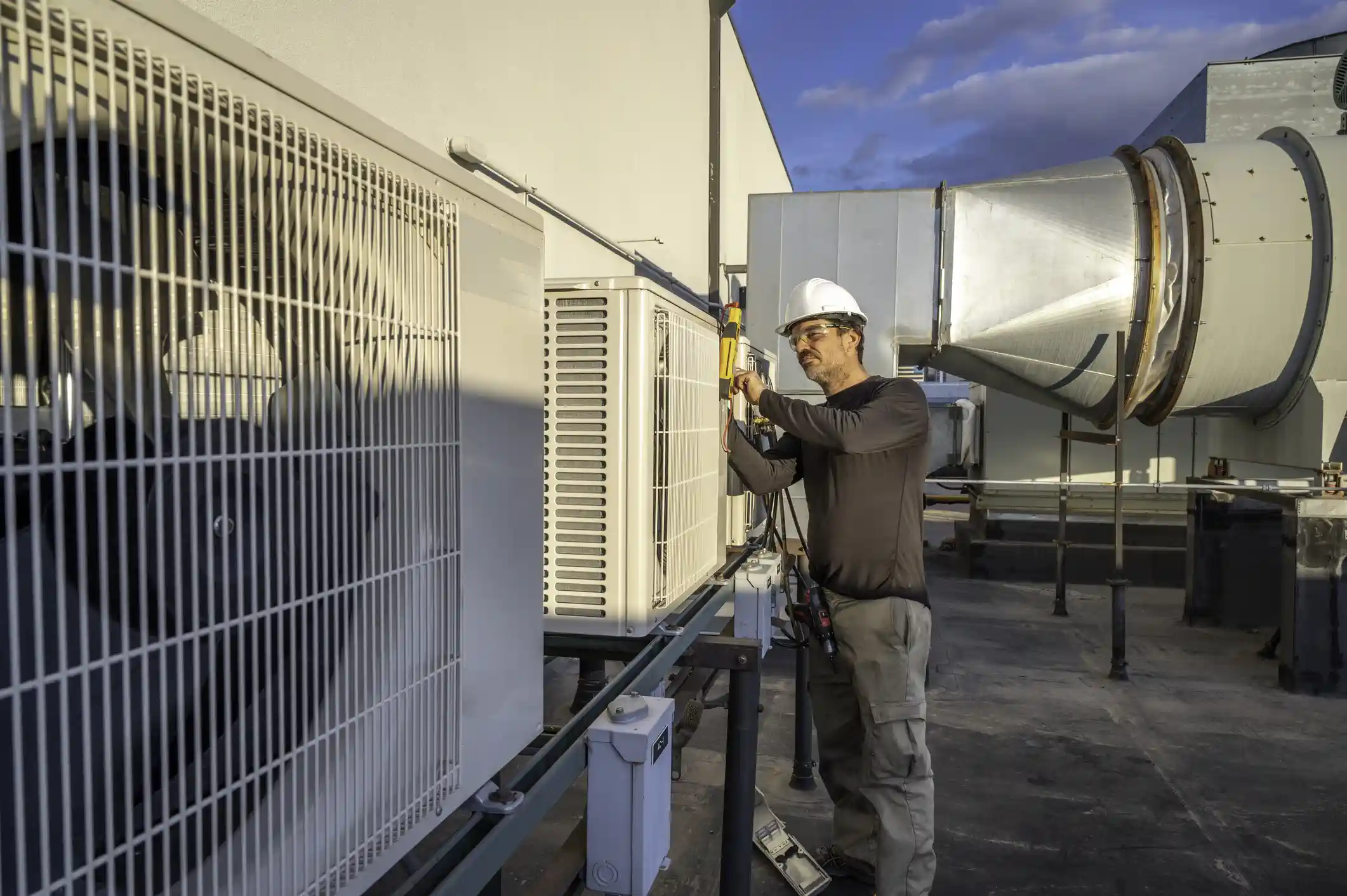 A male HVAC technician wearing a dark shirt, cargo pants, a white hard hat, and safety glasses is working on a row of white commercial air conditioning units installed on a rooftop. He is standing and focused on one of the units, using a tool to work on its side panel. To the right, a large, circular galvanized steel duct extends across the roof. The units are mounted on a metal rack, and the sky above is partly cloudy. The scene depicts professional maintenance on a commercial HVAC system.