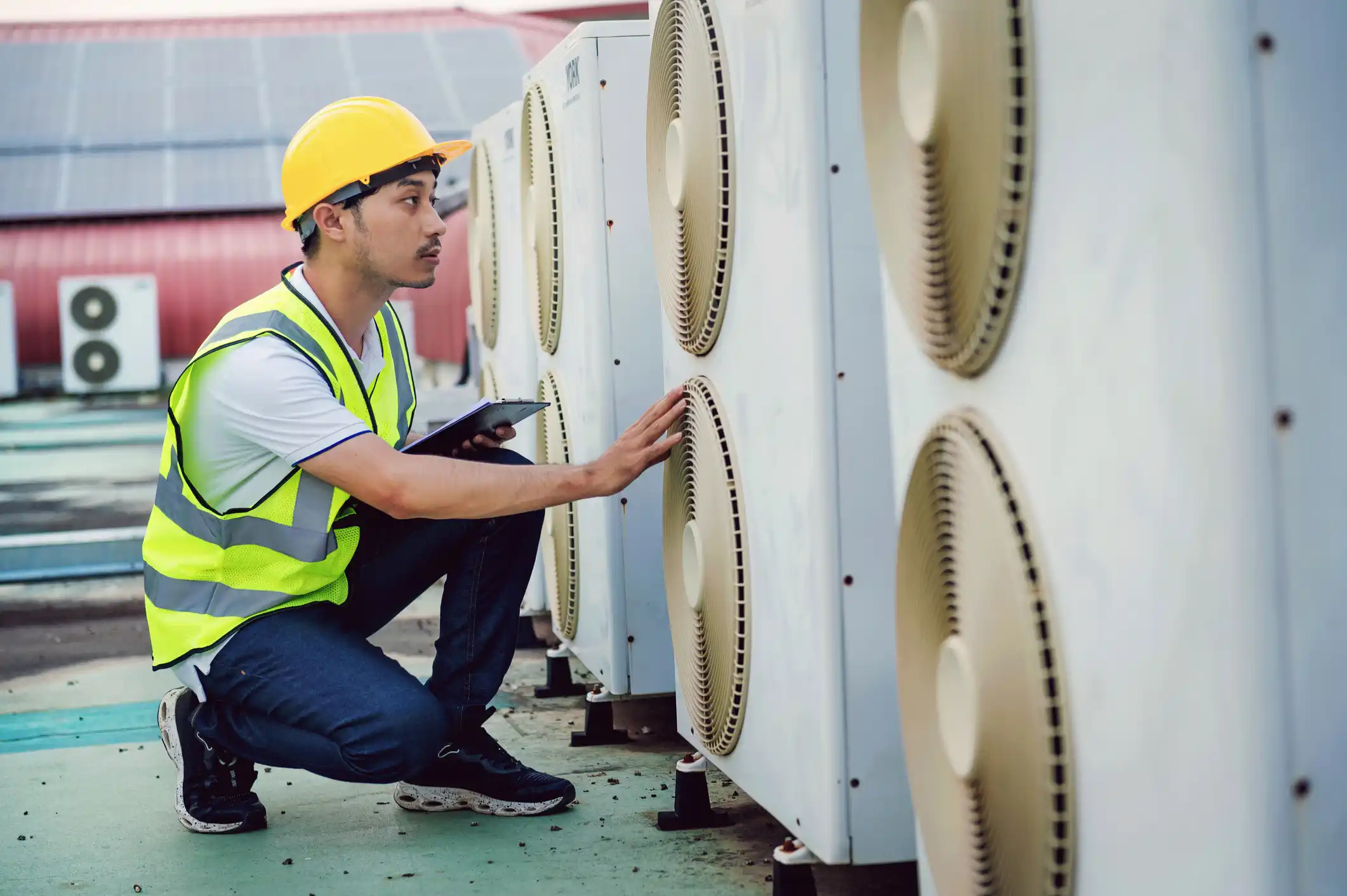 A HVAC technician wearing a white shirt, dark jeans, a bright neon-yellow safety vest, and a yellow hard hat is crouching to inspect a row of white outdoor air conditioning units on a rooftop. He is looking intently at the units, with his right hand resting on the fan grille of one unit while holding a clipboard and pen in his left hand. The background shows a large, flat rooftop with a bank of solar panels and another AC unit visible on the left, suggesting he is performing a professional check-up or maintenance on a commercial HVAC system.