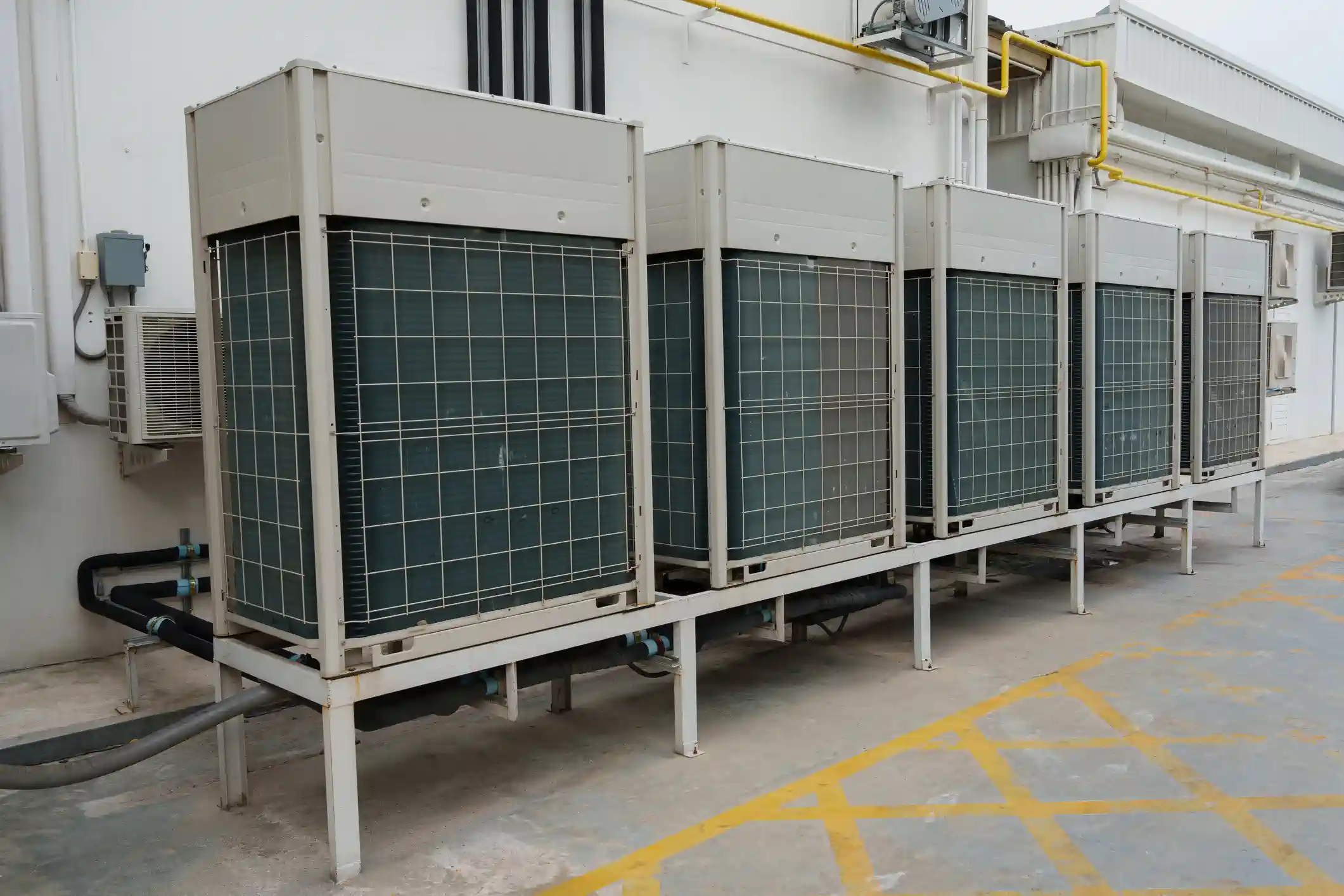 A row of five large, gray commercial air conditioning units (likely outdoor condenser units) is installed on a metal platform against a plain white exterior building wall. Each unit has a dark, gridded grille covering the coils. The platform holds the units off the paved ground, which has yellow safety lines and markings. Various pipes and conduits run along the wall and beneath the platform, including a noticeable yellow pipe near the top right and black insulated pipes near the unit on the left. The scene suggests an industrial or commercial HVAC installation.