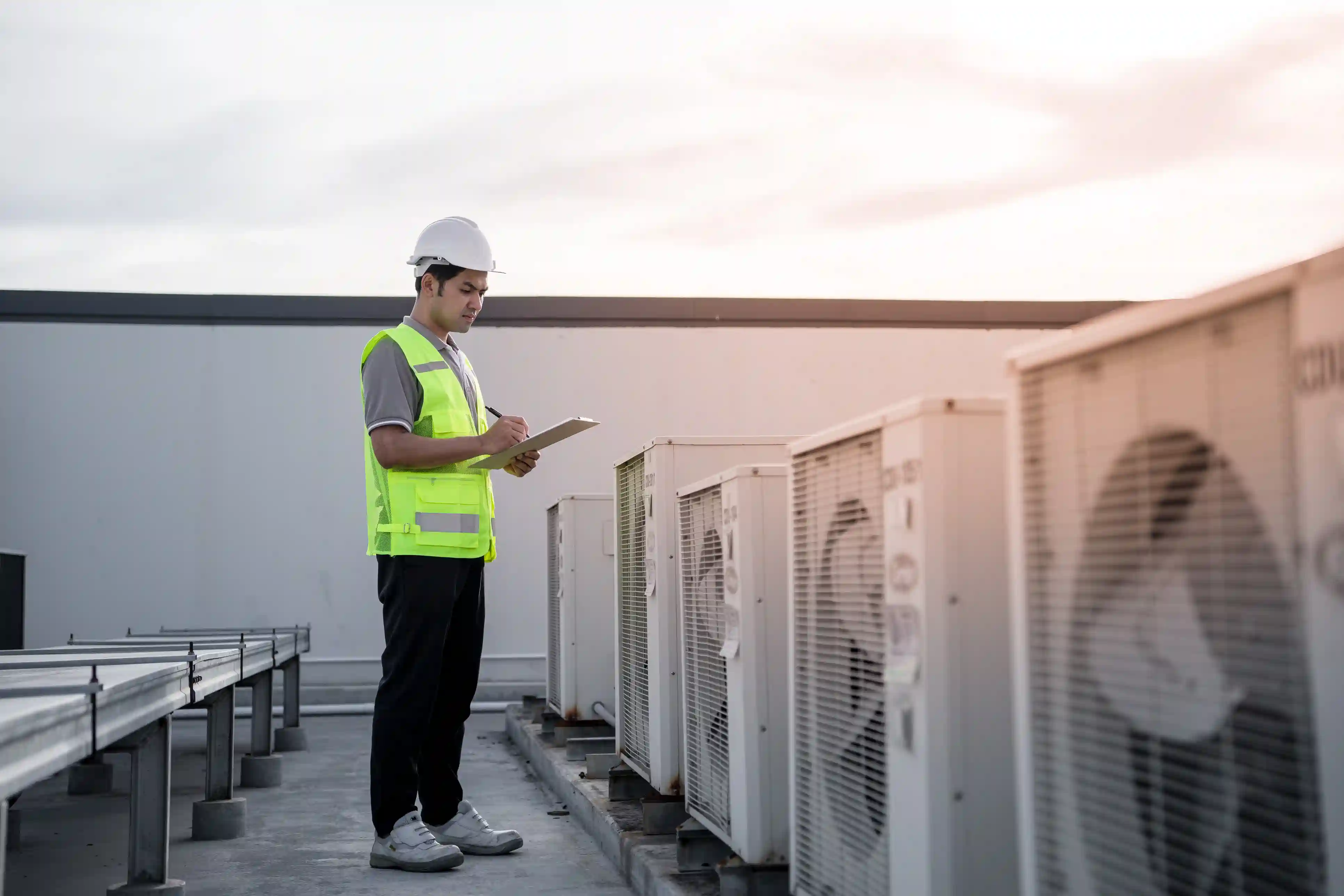 A male HVAC engineer or technician is standing on a rooftop performing an inspection of a row of commercial air conditioning units. He is wearing a white hard hat, a gray polo shirt, black pants, and a bright neon-yellow safety vest. He is holding a clipboard and a pen, writing down notes or checking off items during his inspection. The white AC units are lined up on the right, receding into the distance, with some sort of metal railing or rack visible on the left. The scene is set outdoors under a bright, overcast sky, suggesting an early morning or late afternoon light.