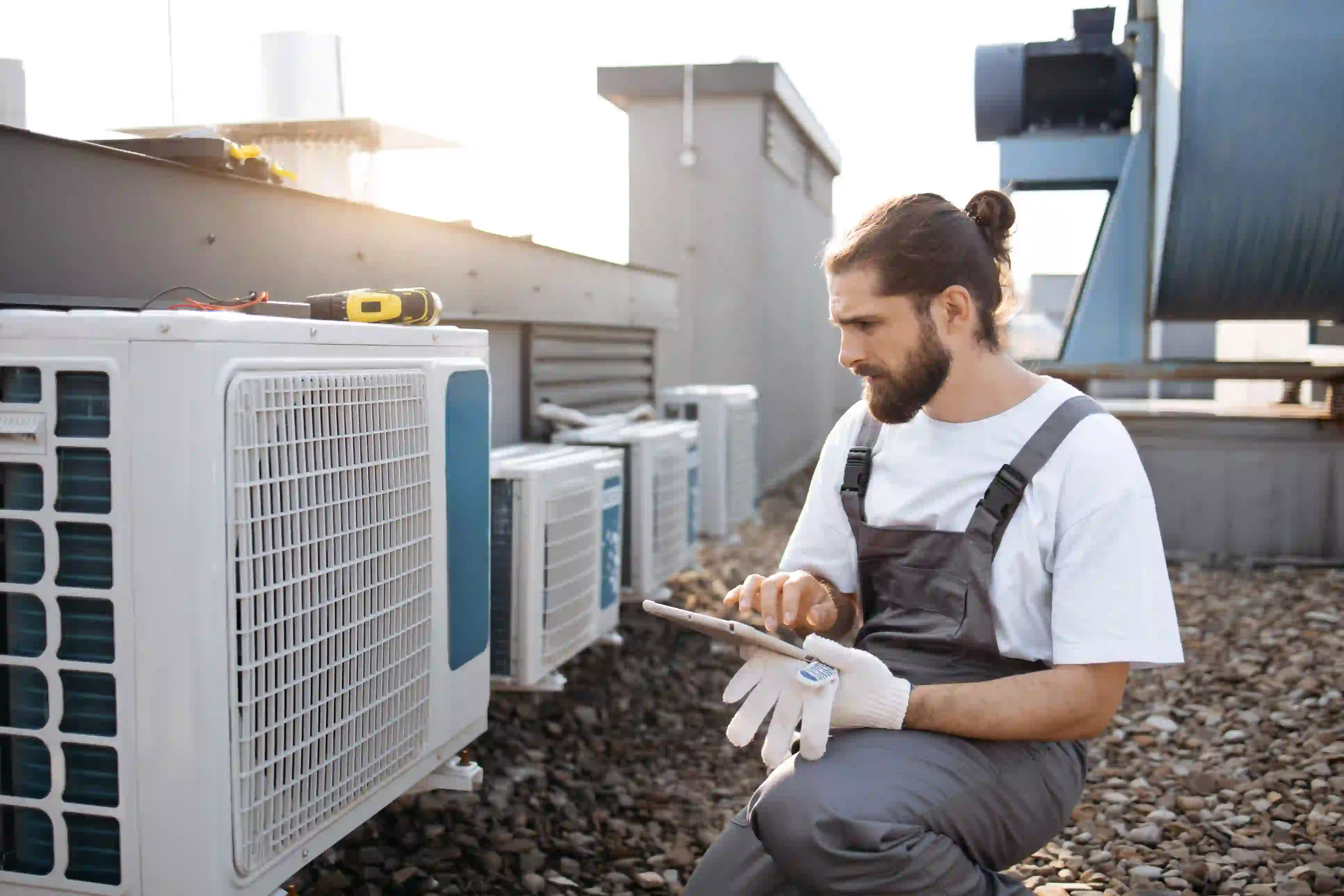 A male HVAC technician with a beard and his hair tied back in a bun is crouching on a gravel-covered rooftop to inspect a row of white air conditioning units. He is wearing a white shirt and gray overalls, with a white work glove in his lap. He is using a digital tablet with his right hand, likely checking diagnostics or logging maintenance data. Several AC units are visible along a wall, suggesting a commercial installation, with large ventilation equipment in the background and a bright, sunny sky with lens flare on the left.