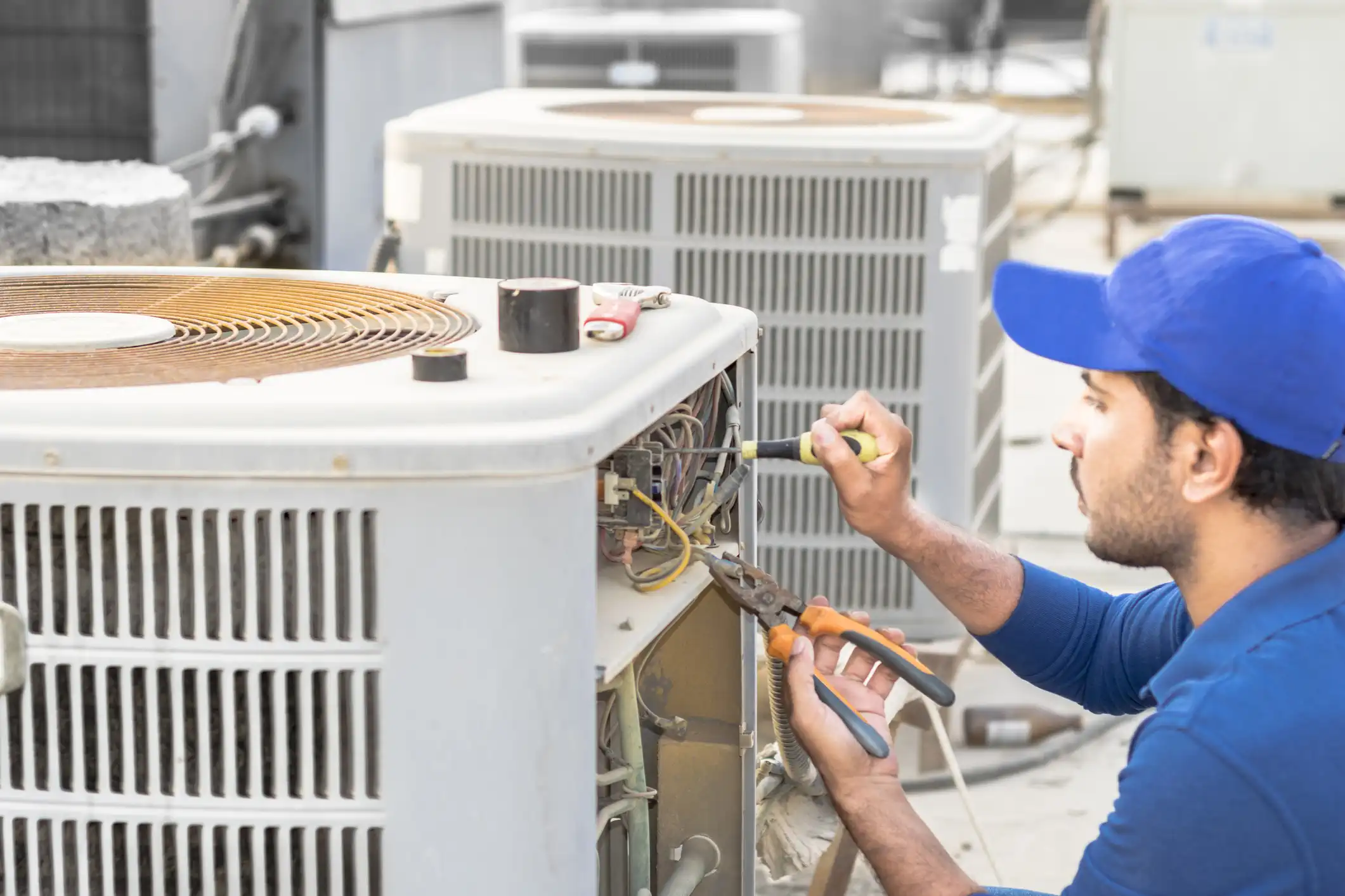 A male HVAC technician wearing a bright blue baseball cap and a matching blue uniform shirt is focused on performing repair or maintenance work on a large, white outdoor air conditioning condenser unit. He is using pliers with orange handles in his left hand and a screwdriver with a yellow handle in his right hand to access the electrical wiring inside the unit's open compartment. The work is taking place on a rooftop, with another gray AC unit visible in the blurred background. A small roll of black electrical tape is sitting on top of the white unit.
