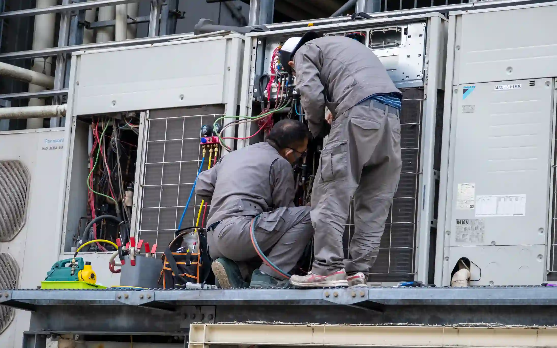Two HVAC technicians wearing gray work jumpsuits are working on a bank of large, commercial Variable Refrigerant Flow (VRF) air conditioning units on a metal platform. Both units have their service panels open, revealing extensive wiring, piping, and components. One technician is kneeling, intently working on the lower section, while the other is standing and bent over the upper part of the unit. Various tools and specialized equipment, including a yellow and green refrigeration recovery or vacuum pump and a tool bag, are visible on the platform, indicating a major repair or installation of a complex commercial HVAC system.