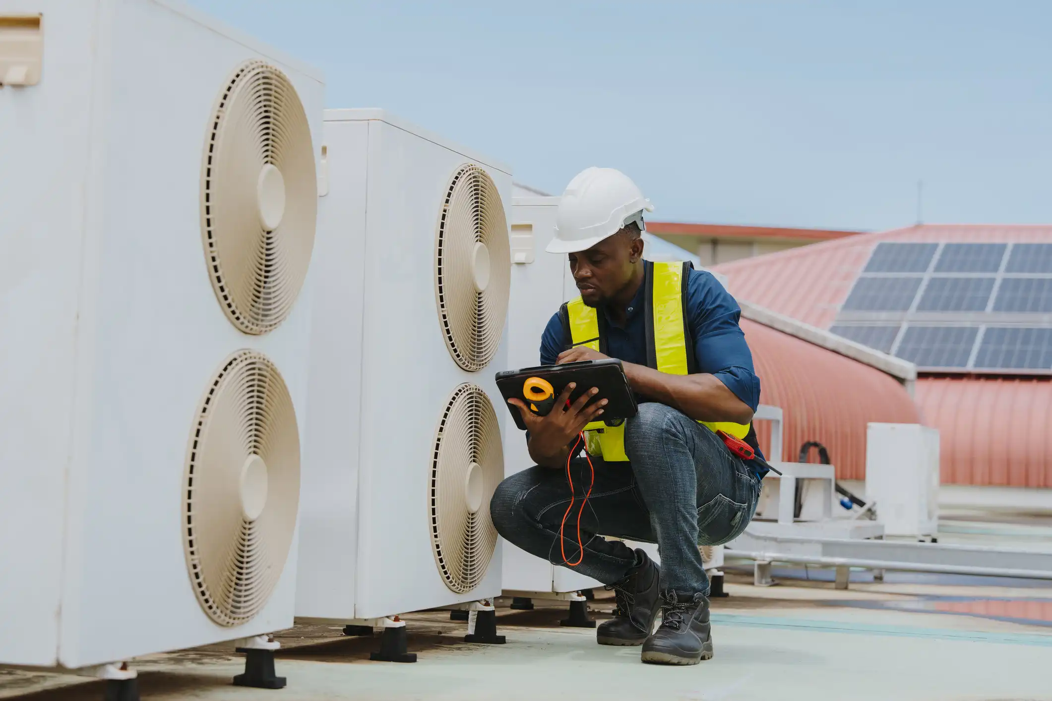 A Black male HVAC technician is crouching on a rooftop to inspect a bank of large, white commercial air conditioning units. He is wearing a white hard hat, a blue shirt, jeans, a neon-yellow safety vest, and work boots. The technician is focused on a digital tablet he holds with both hands, likely using it to run diagnostics, record performance data, or access service manuals. The AC units feature two prominent circular fan grilles each. In the background, a row of solar panels is visible on a lower, reddish-colored roof, suggesting the technician is maintaining a modern, energy-conscious facility.