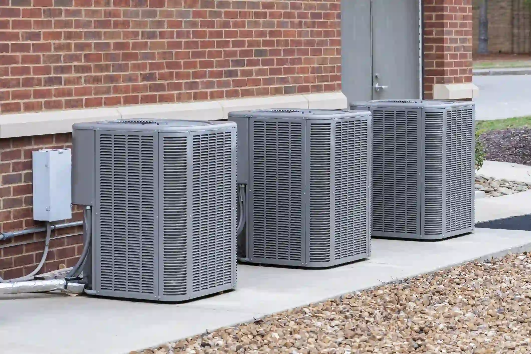 Three large, gray outdoor air conditioning condenser units are lined up on a concrete walkway next to a building with an exposed red brick wall. The units are uniformly sized, featuring horizontal louvers on all sides. An electrical disconnect box is mounted on the wall next to the leftmost unit, with conduit and insulated piping visible near the base. The area in front of the walkway is covered in light-colored gravel, and a gray door is visible on the building to the right of the AC units.
