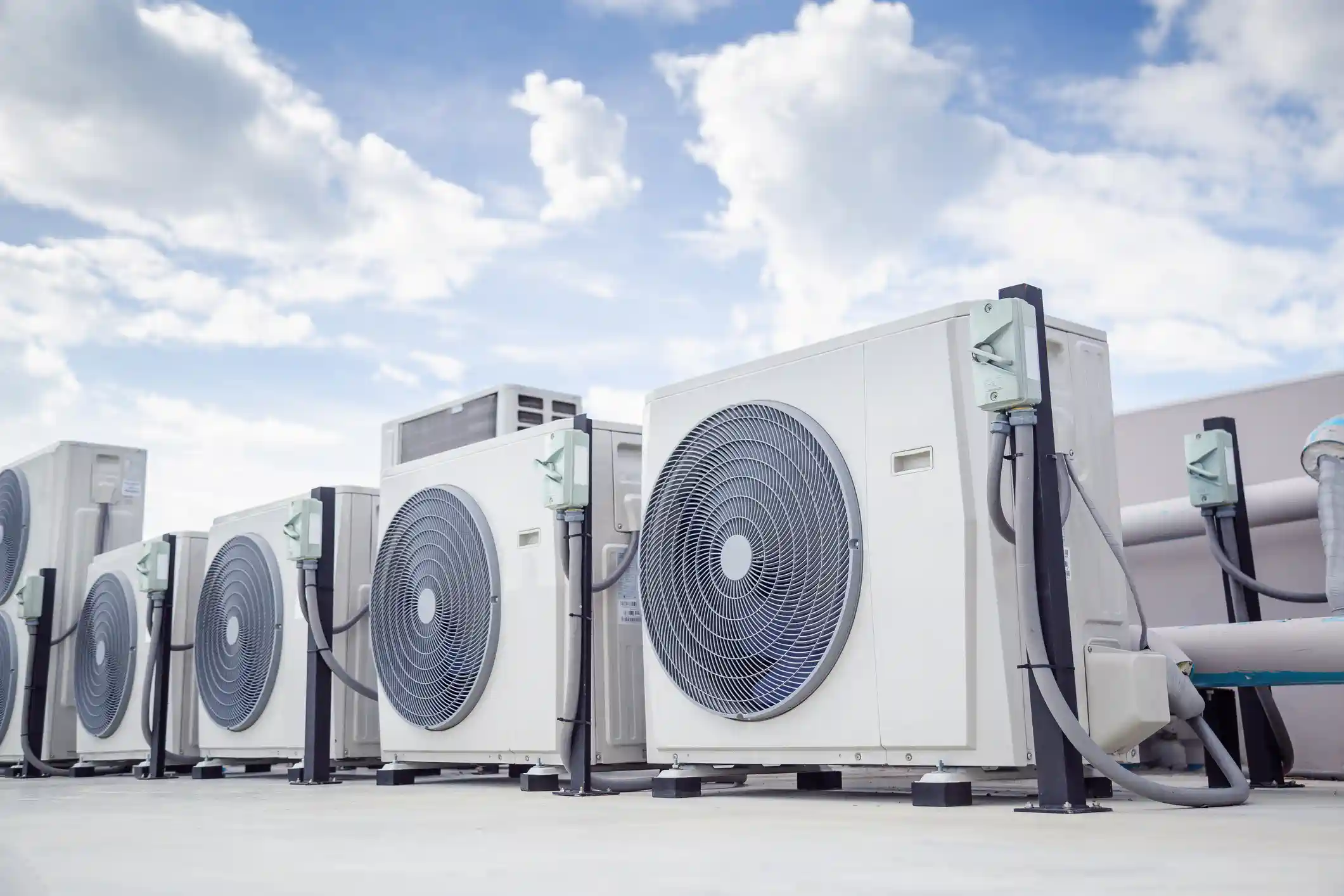 A low-angle view shows a long row of white outdoor air conditioning condenser units installed on a flat, paved rooftop. The units are lined up and feature large, circular fan grilles. Each unit is connected to a black vertical service column with a mounted electrical disconnect switch. The entire installation is set against a bright, blue sky filled with white clouds, emphasizing a large-scale commercial HVAC system.