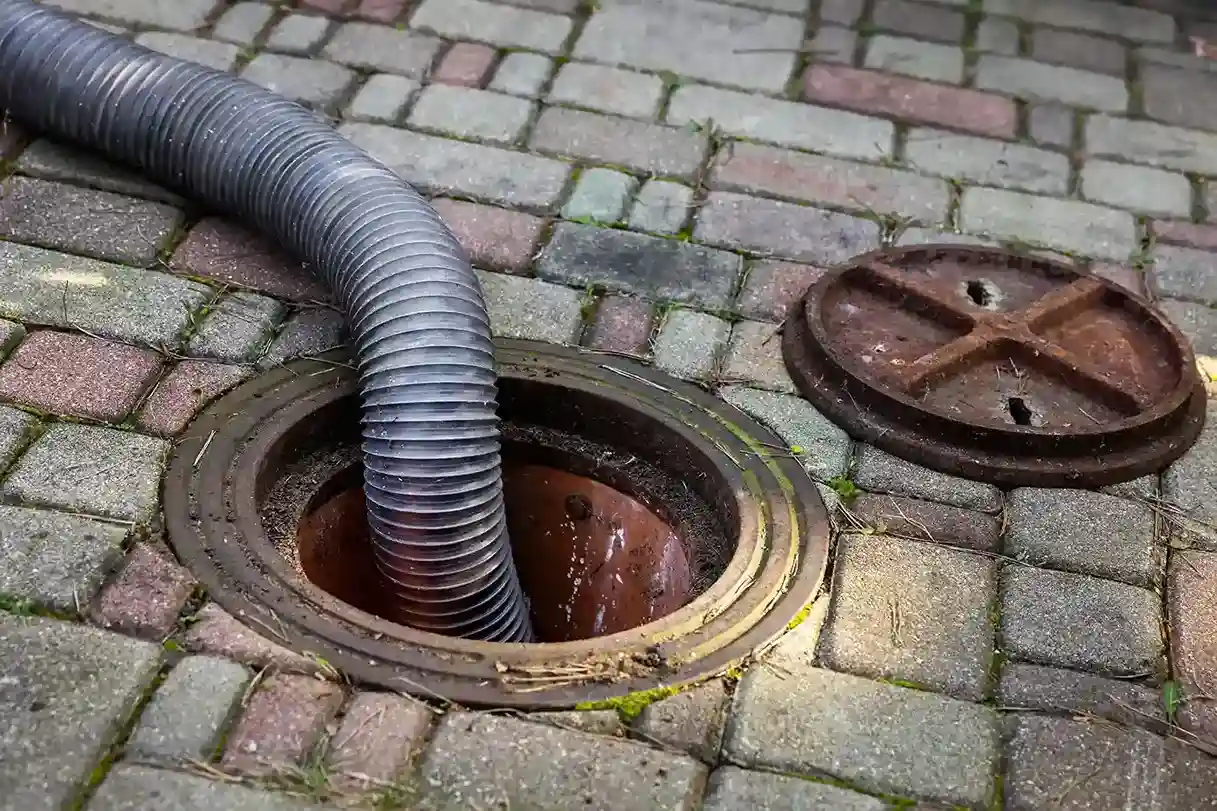 Close-up of a large corrugated hose inserted into an underground inspection chamber or septic access on a brick patio.