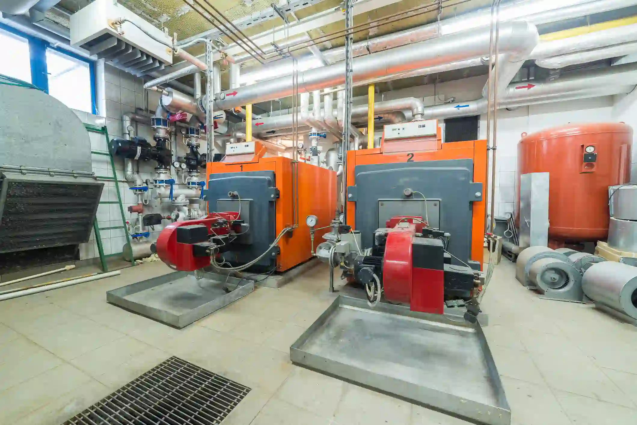 An industrial or large commercial boiler room featuring two identical, large, orange, and dark grey boilers (heating units), numbered 1 and 2. Each boiler has a prominent red and black burner unit attached and is sitting in a metal drip pan. The room is filled with extensive overhead metal and insulated piping, an orange expansion tank on the right, and various other components like metal ductwork and fans on the floor. A floor grating is visible in the foreground, and there is a window providing daylight on the upper left. This represents a heavy-duty heating system installation.