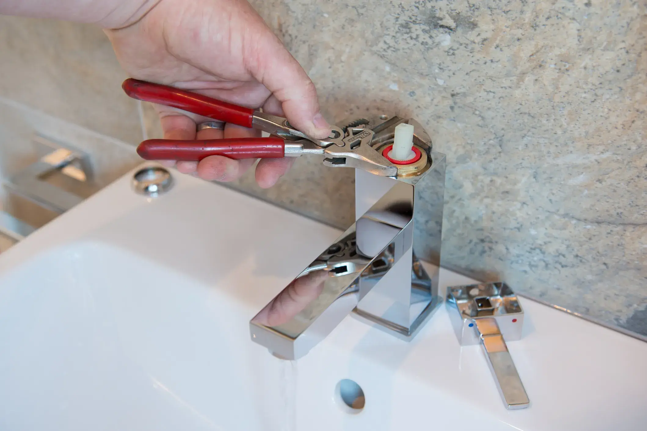 Close-up of a hand using channel lock pliers to remove the valve cartridge for repair from a modern bathroom faucet.