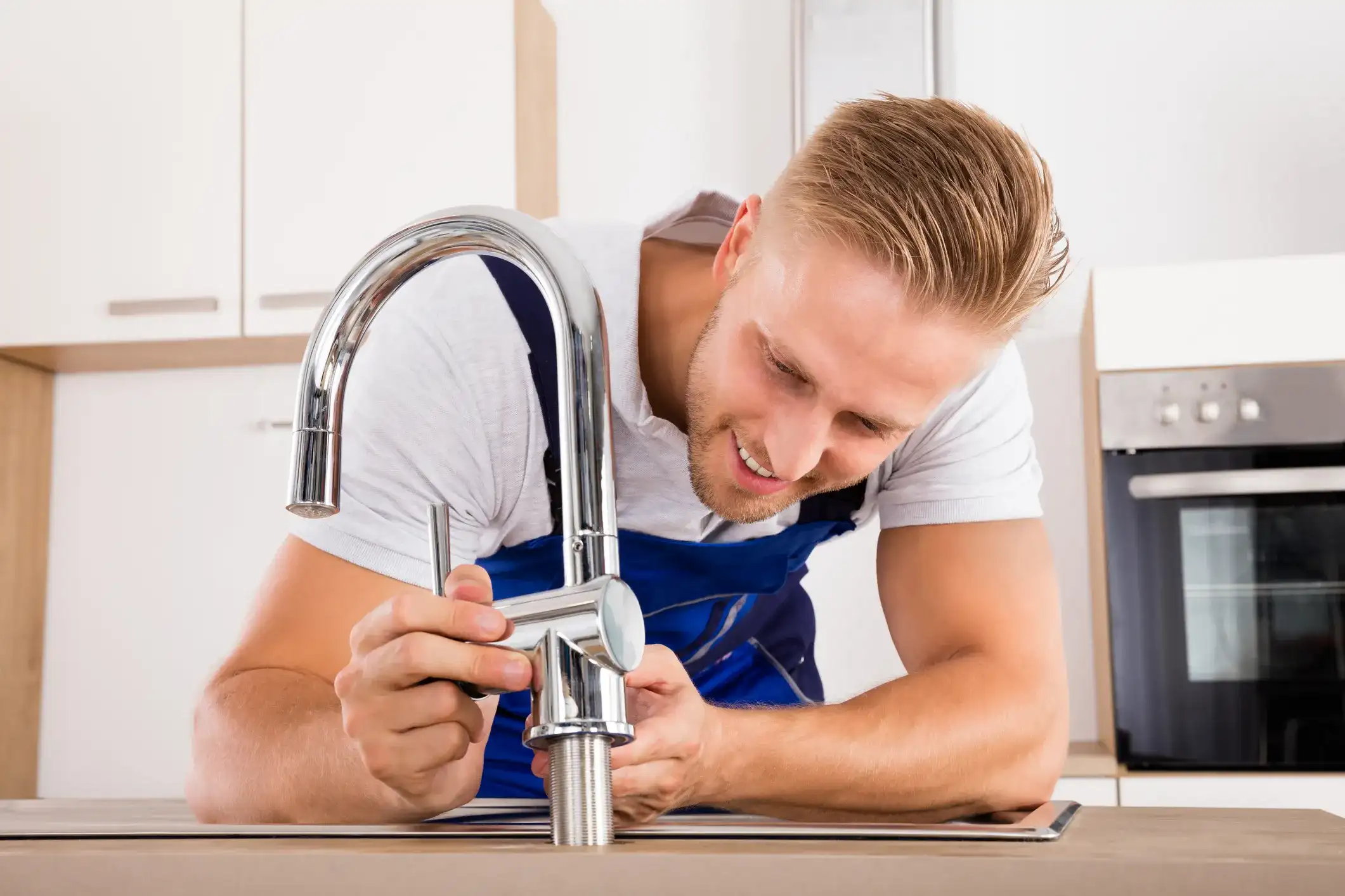 Professional plumber in blue uniform installing or repairing a new kitchen faucet on a countertop with small tile backsplash.
