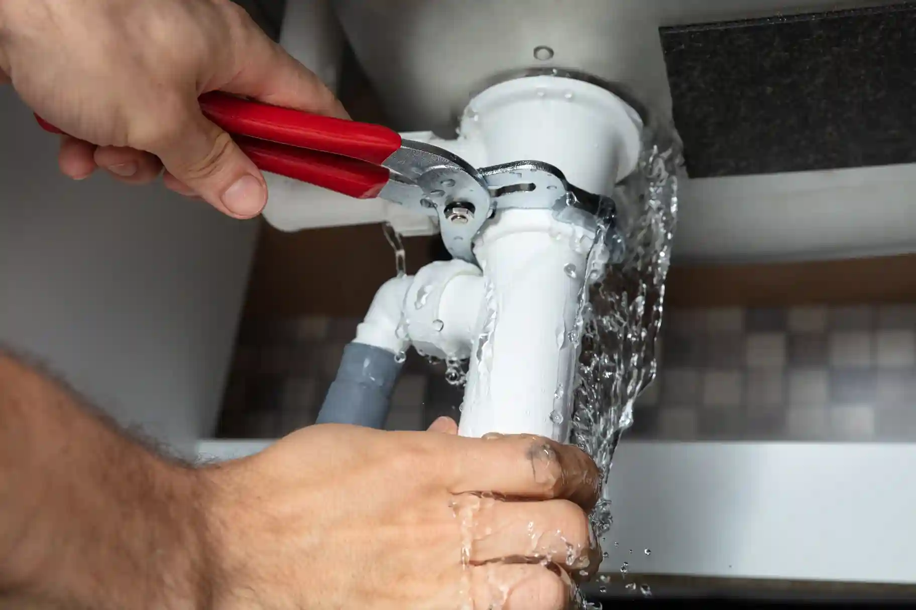 Close-up of a plumber using channel lock pliers to repair a burst or leaking pipe connection under a sink, with water spraying out.