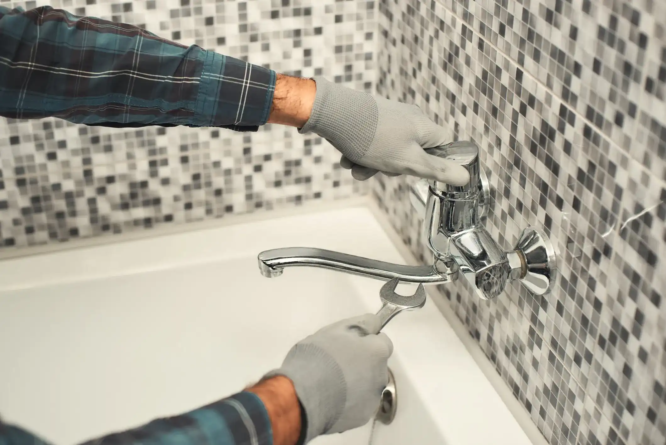 Plumber in gloves using a wrench to repair a bathtub mixer faucet against a mosaic tile bathroom wall.