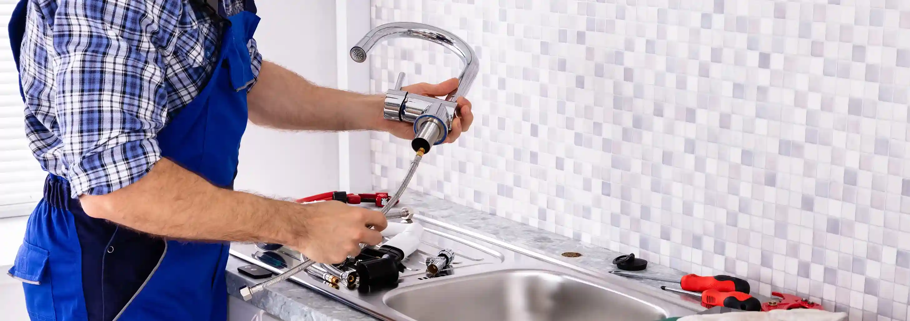 Professional plumber in overalls preparing to install a new kitchen faucet above a stainless steel sink with a tiled backsplash.