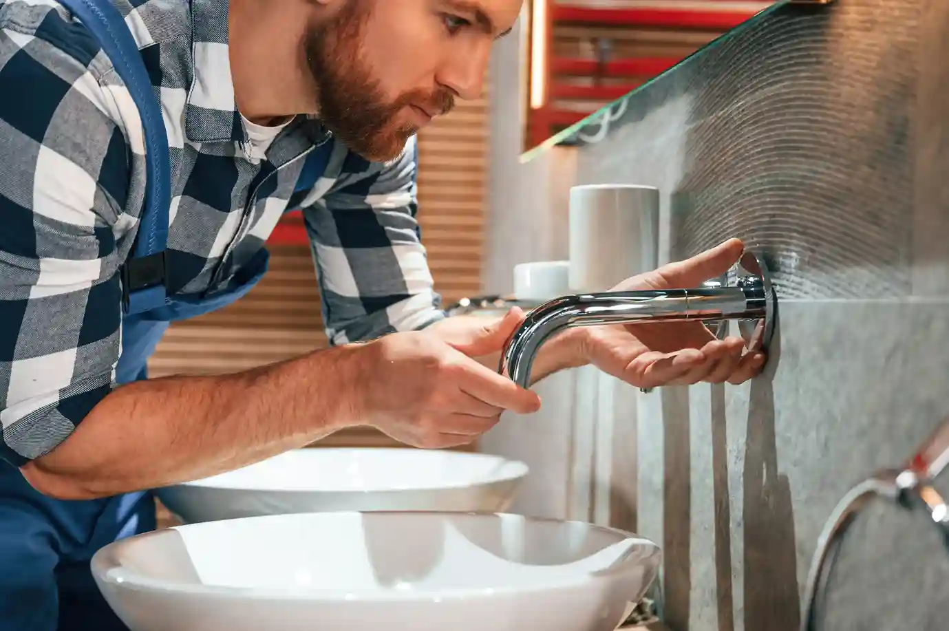 Plumber installing or adjusting a wall-mounted chrome faucet over two modern vessel sinks in a bathroom.