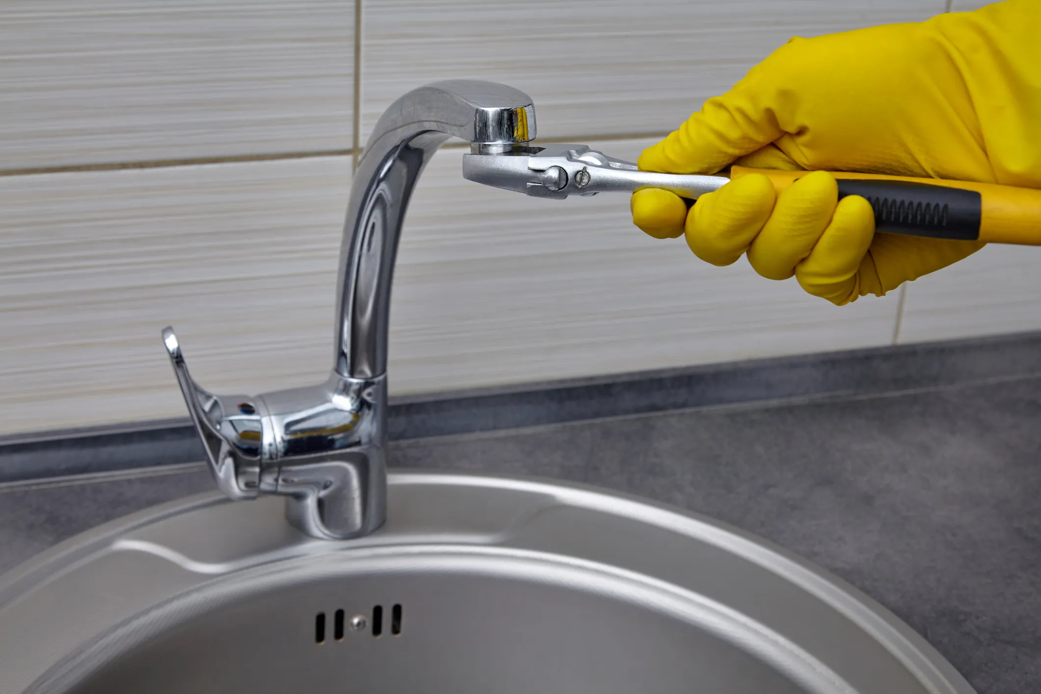 Hand in a yellow glove using an adjustable wrench to tighten the aerator or spout of a chrome kitchen faucet over a stainless steel sink.
