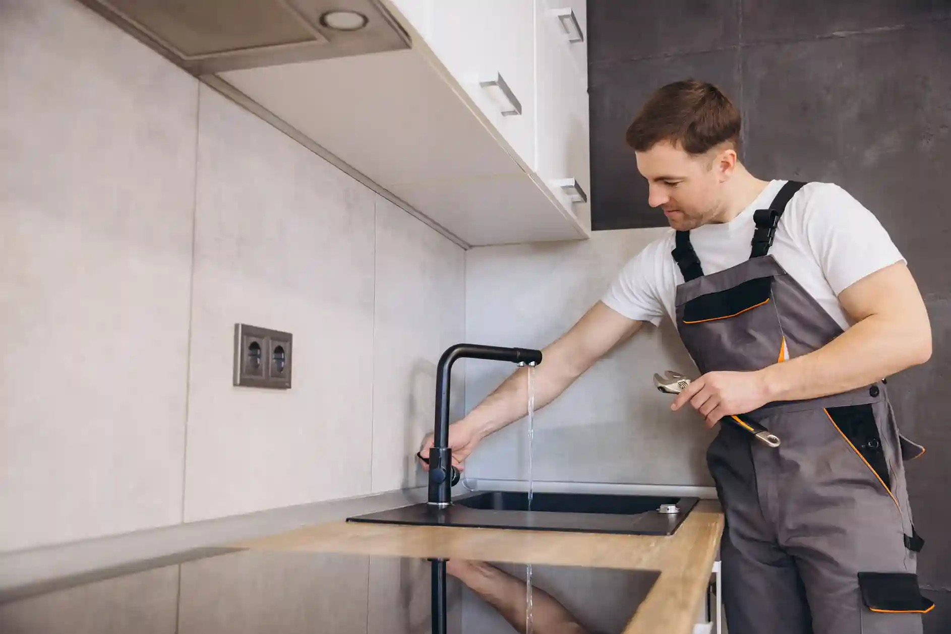 Professional plumber in overalls checking the water flow of a new matte black kitchen faucet after installation and holding a wrench.