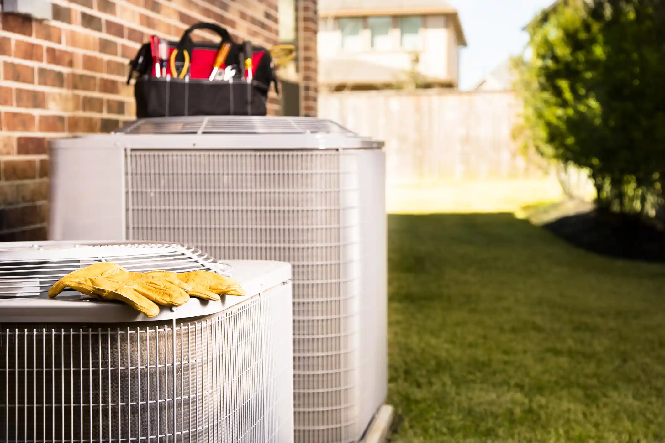 Two large, light-grey outdoor air conditioning or heat pump units are positioned next to a red brick building on a sunny day. A pair of yellow work gloves is resting on the grille of the unit in the foreground. A black and red tool bag, filled with assorted tools, is visible on top of the rear unit. The units are set on a concrete pad, with a well-maintained lawn and wooden fence in the background, suggesting a residential HVAC service scenario.