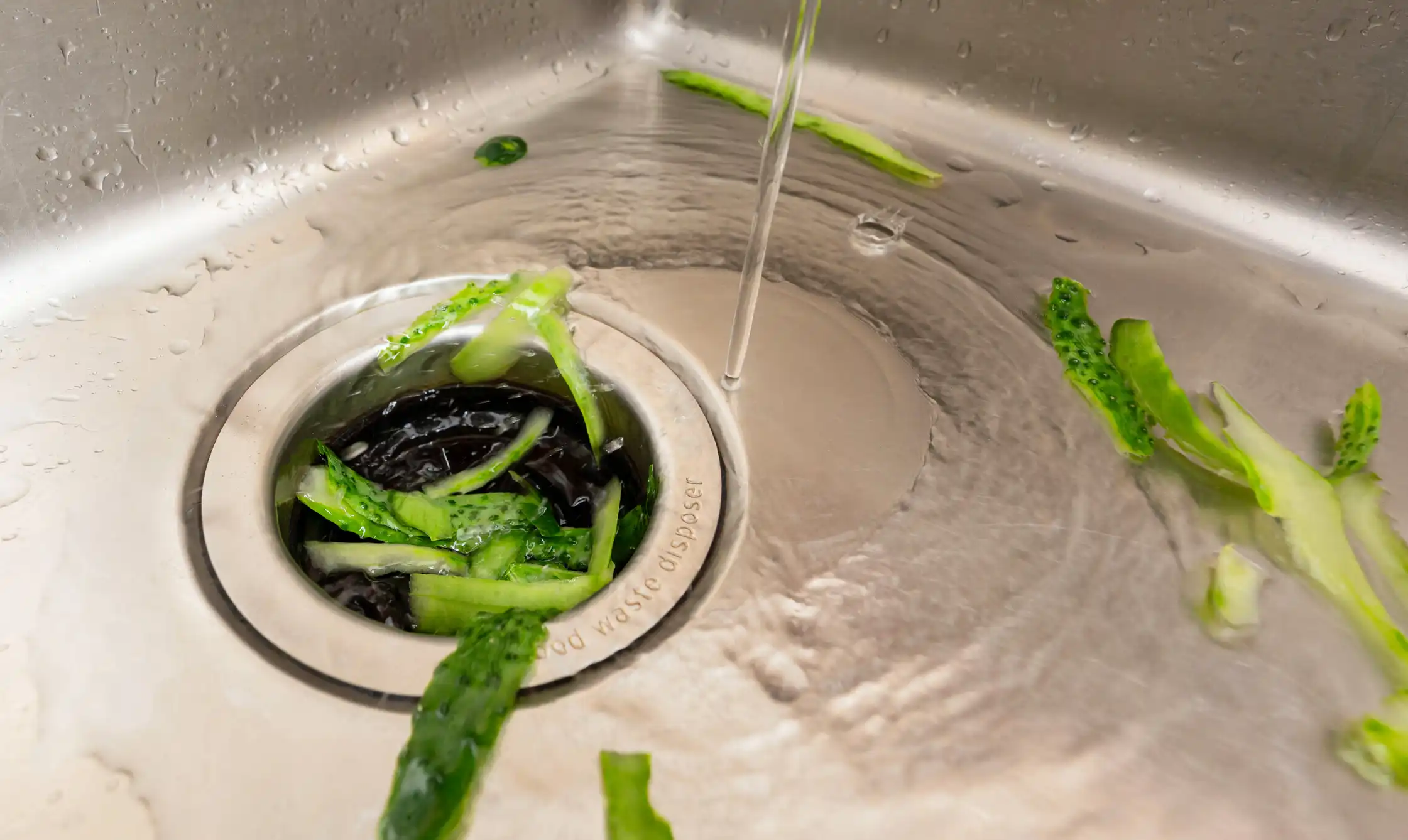 Cucumber peels and running water going down into a kitchen sink's food waste disposer.