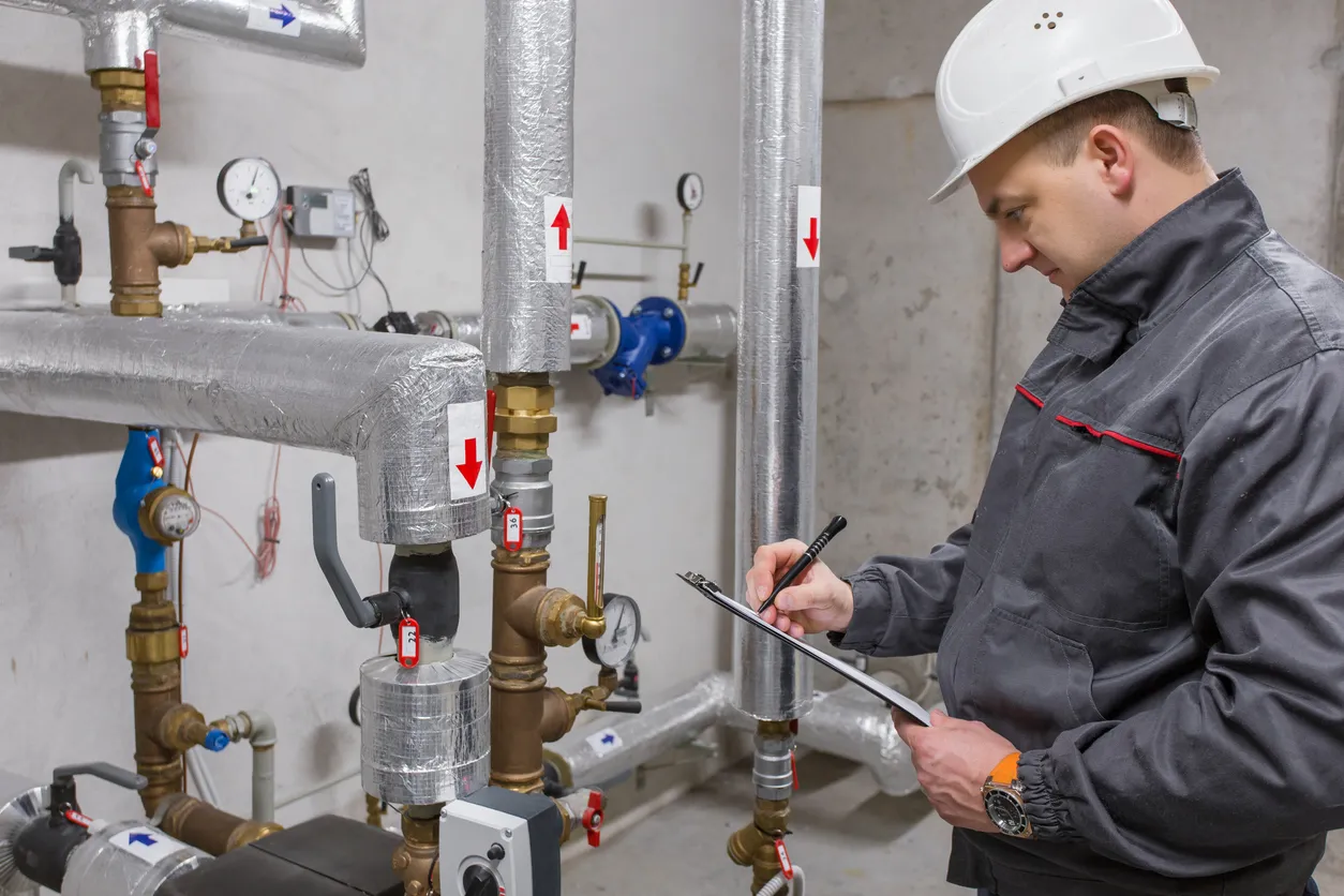 An inspector in a hard hat and gray jacket examines industrial pipes with gauges, writing notes on a clipboard.
