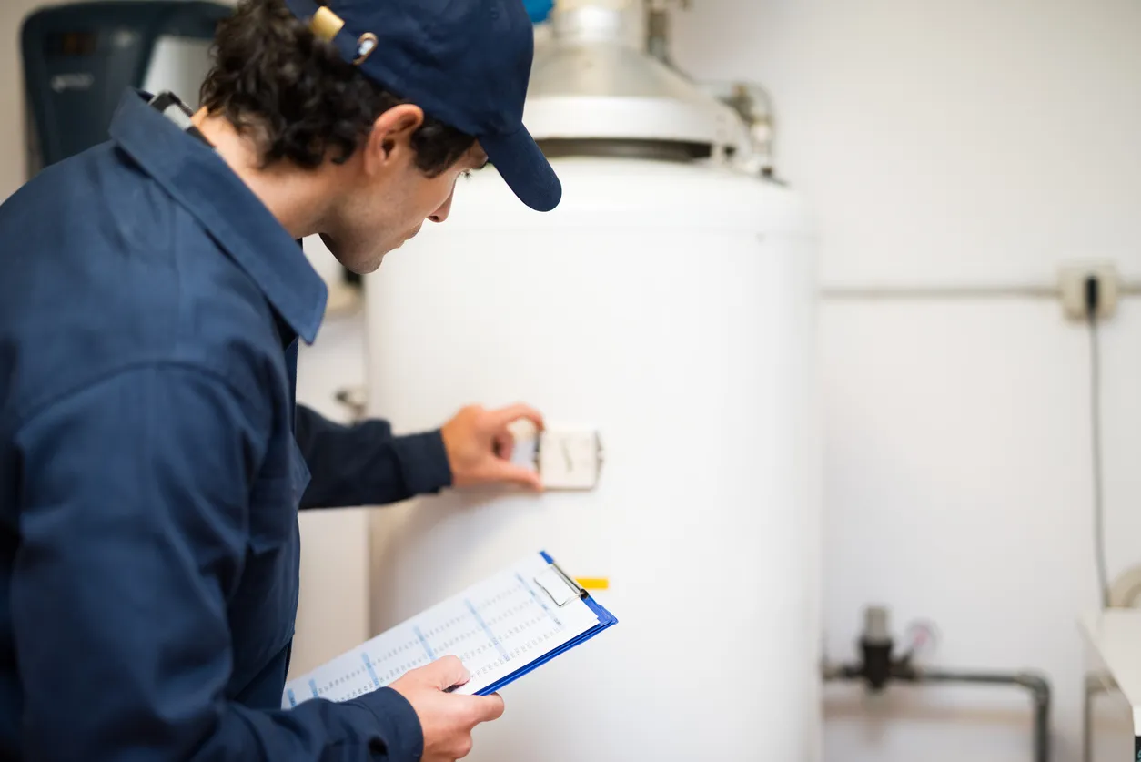 A technician in a blue cap inspects the control panel of a large white water heater, holding a clipboard.