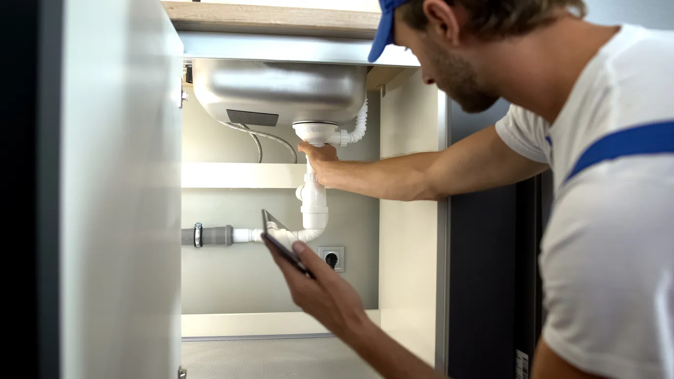 A plumber in a white shirt inspects drain pipes under a kitchen sink, holding a smartphone.