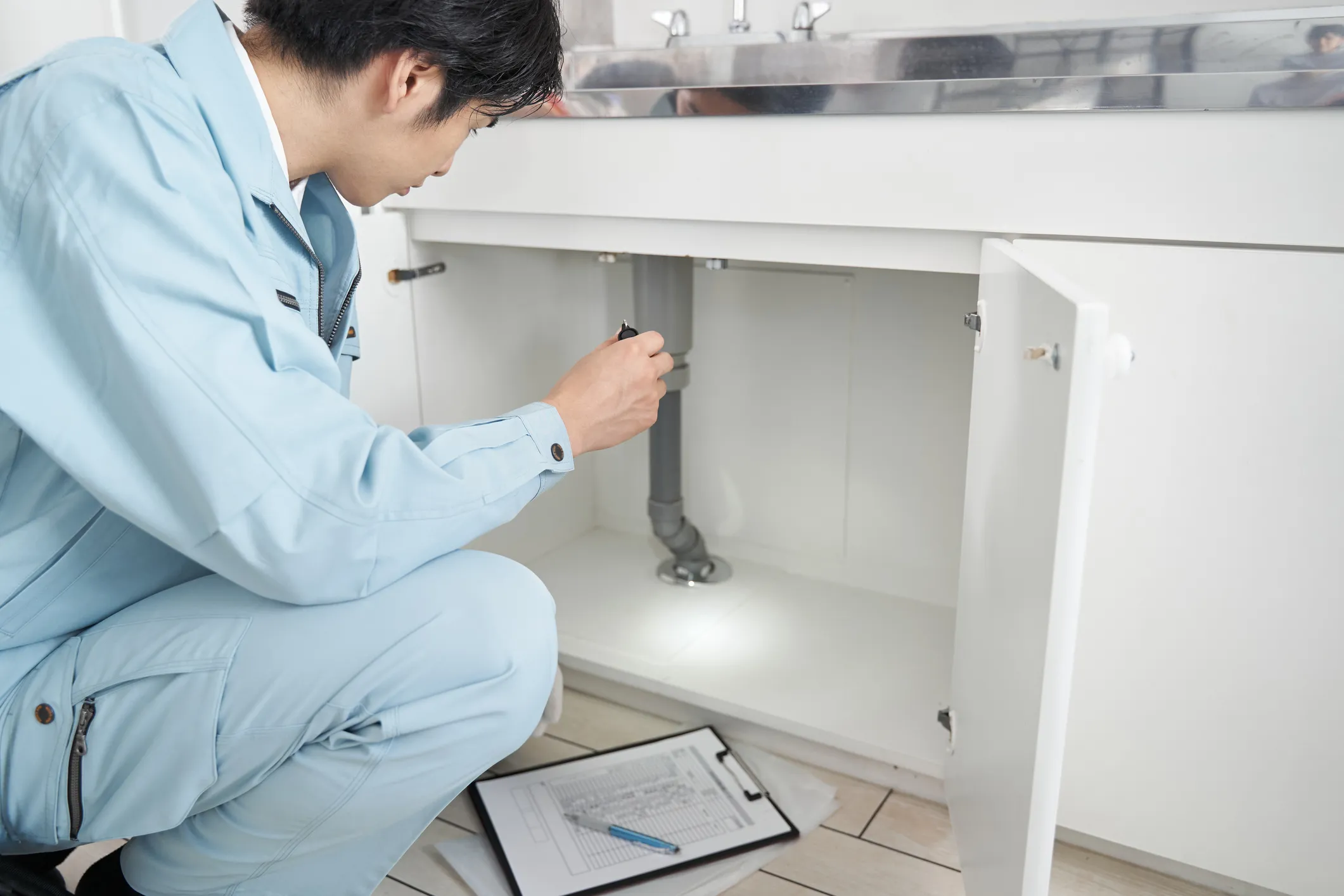 A technician in a light blue jumpsuit crouches to inspect pipes under a white cabinet, using a flashlight.