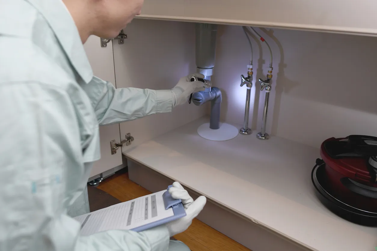 A technician in gloves and a light green shirt inspects the drain trap and pipes under a cabinet, holding a clipboard.