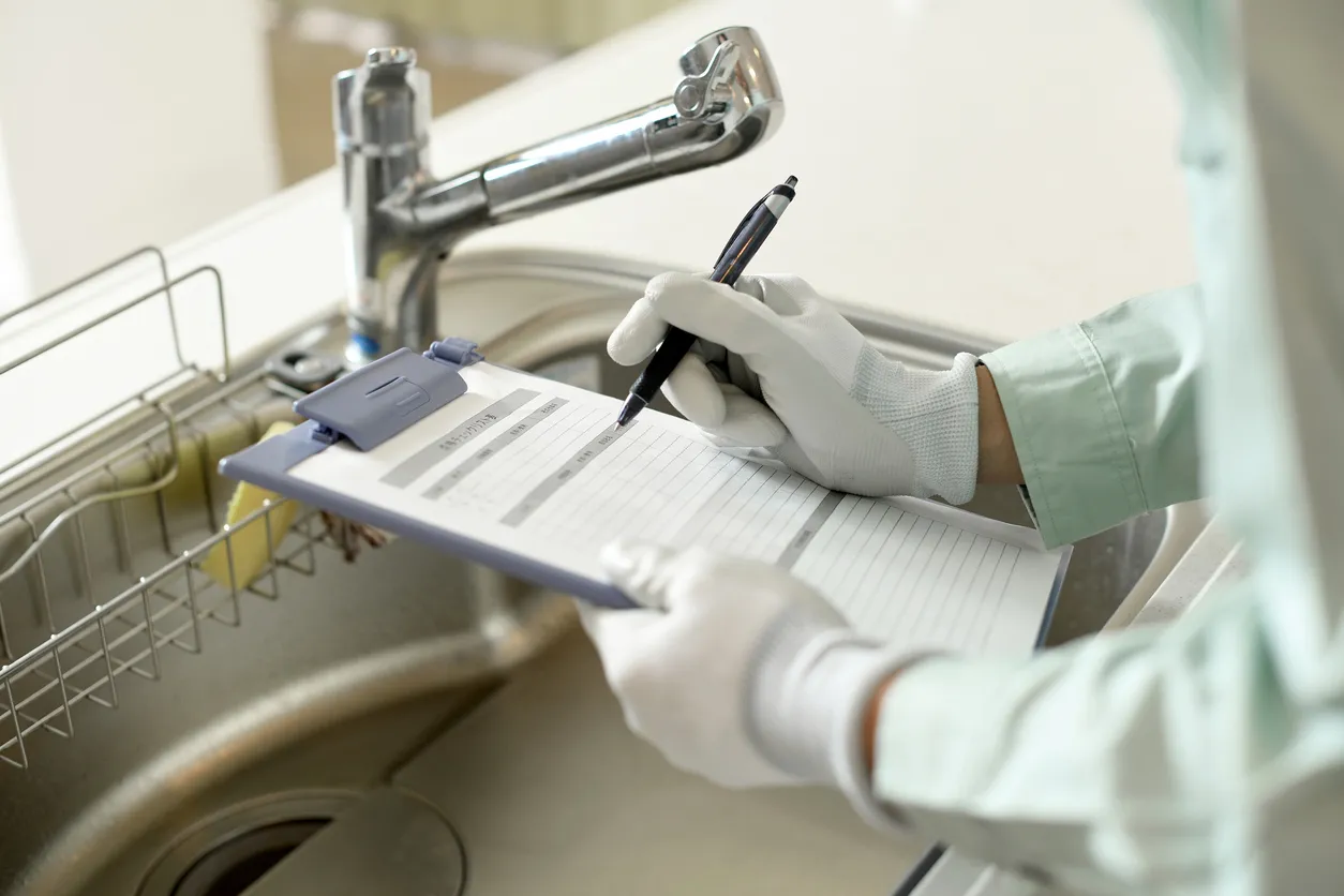 Close-up of gloved hands filling out an inspection form on a clipboard next to a kitchen sink faucet.