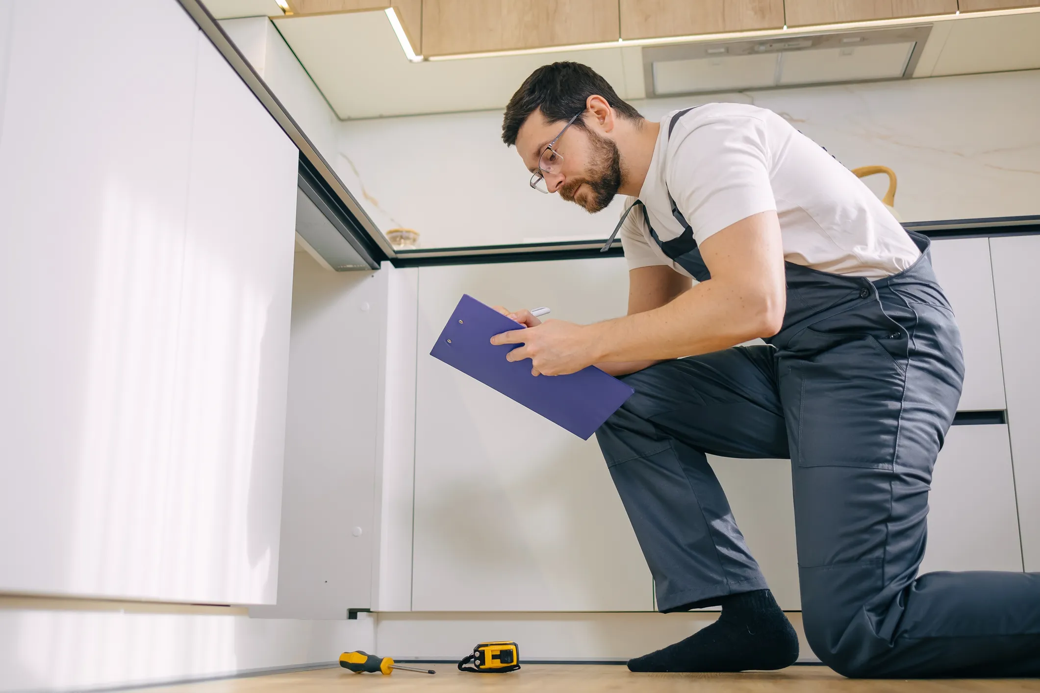 A plumber in glasses and gray overalls kneels in a modern kitchen, writing on a blue clipboard during an inspection.
