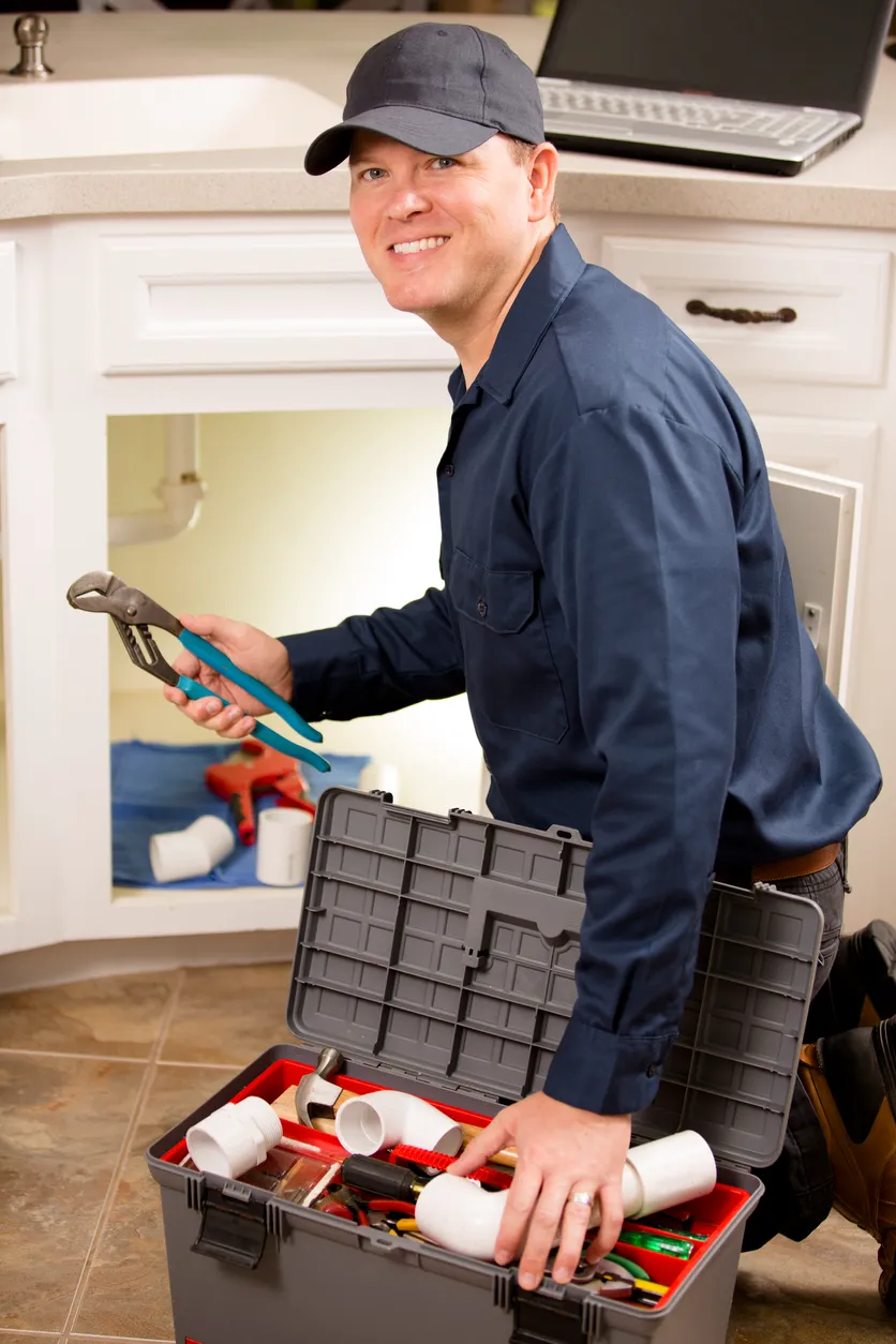 Smiling plumber kneeling with an open toolbox full of plumbing tools and pipes.