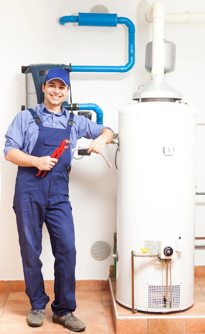 Smiling plumber in overalls holding a wrench next to a large water heater and blue piping.