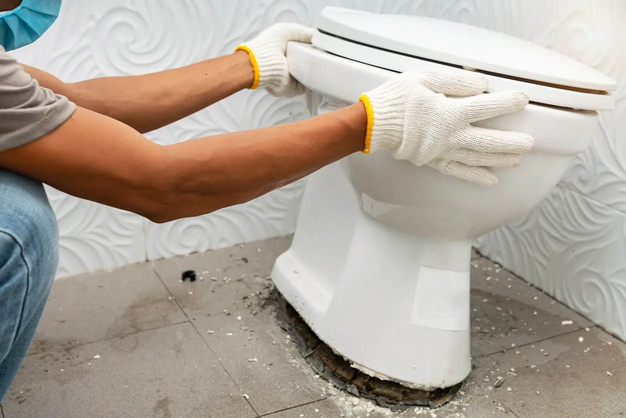 A plumber's gloved hands lift or set a new toilet bowl onto the flange for replacement.