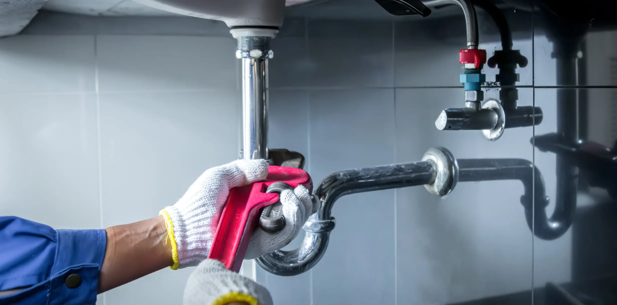 A person wearing a white and yellow-rimmed glove is using a red pipe wrench to tighten a chrome P-trap connection beneath a sink. In the background, there are hot and cold water valves with red and blue accents, all set against a dark gray tiled wall.
