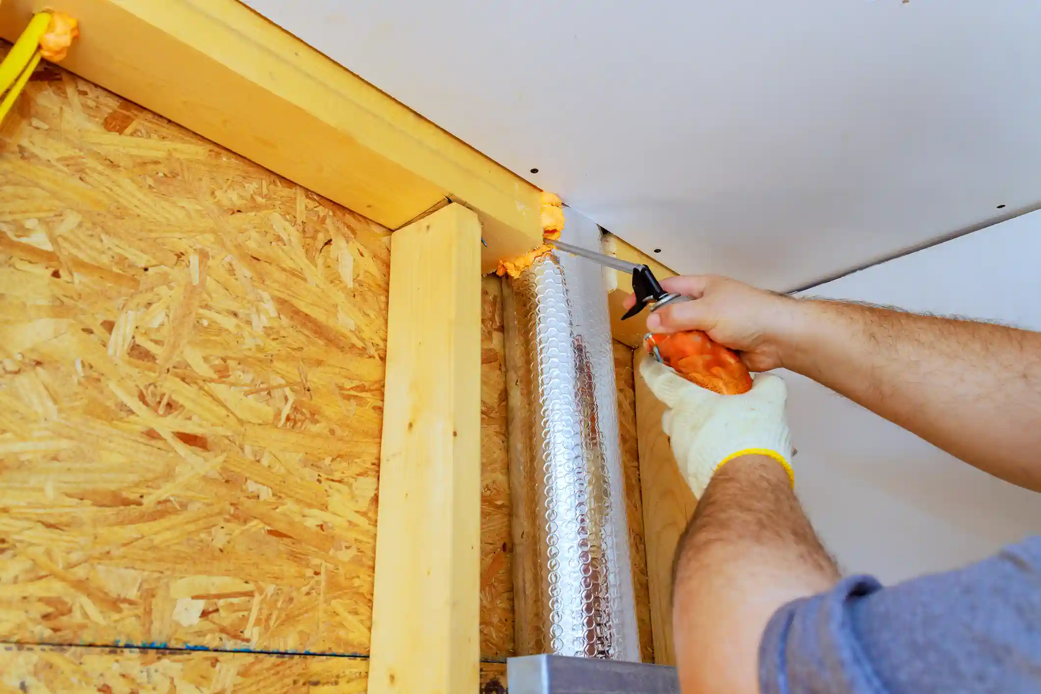 A close-up shot shows a worker's hand, wearing a white glove, using a caulk gun to apply orange expanding foam sealant to the gap between a wooden stud frame and a white drywall ceiling/panel. OSB (Oriented Strand Board) sheathing is visible on the wall to the left, and a foil-wrapped duct runs vertically in the center.