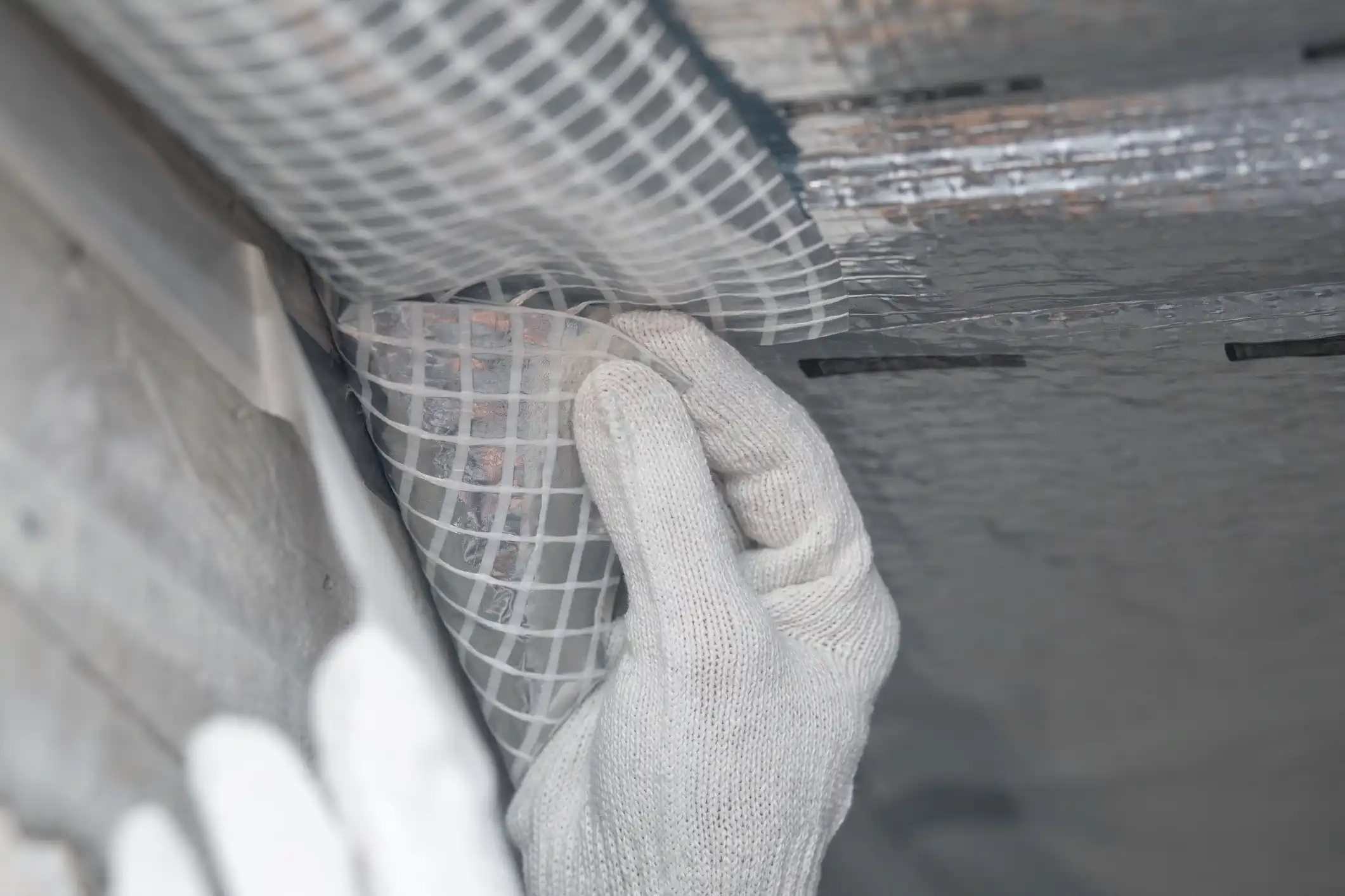  A close-up image shows a gloved hand pressing a piece of white, grid-reinforced material against a reflective, foil-backed surface, likely an insulation product or vapor barrier in a crawl space or attic. The material being applied has a clear, plastic mesh structure.