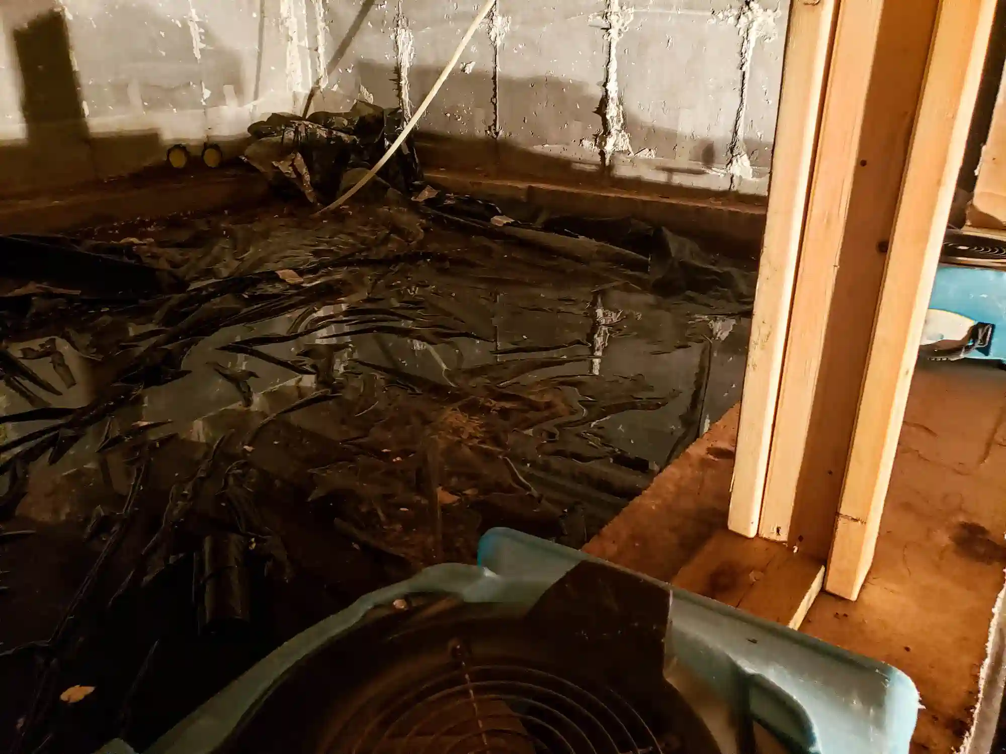 An indoor image of a dirty, unfinished crawl space or basement shows a crumpled black plastic sheet covering the ground, indicating a makeshift vapor barrier. The concrete foundation wall is rough and worn. In the foreground, there are wooden studs and a light blue drying fan/air mover.