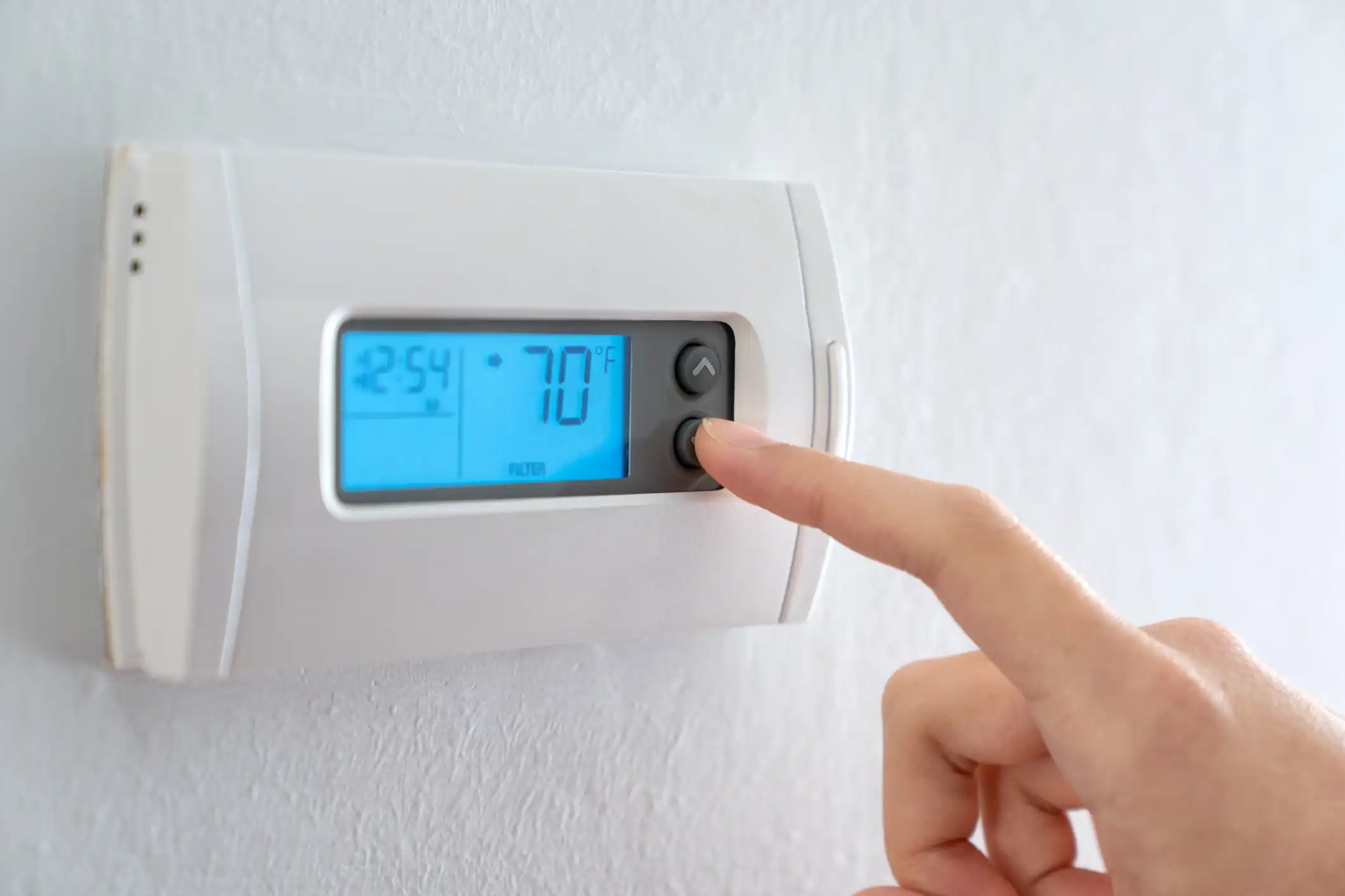 Close-up of a hand pressing a button on a standard white digital thermostat displaying '$77^\circ$' F.