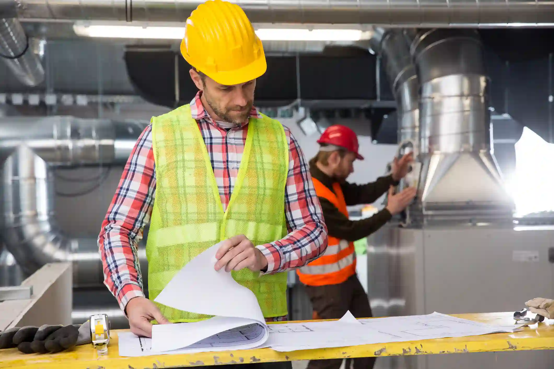  A construction or HVAC manager wearing a yellow hard hat and a high-visibility vest over a plaid shirt is looking down at and reviewing blueprints or technical plans spread out on a table. In the background, another worker in a red hard hat is adjusting a large metal ductwork component.