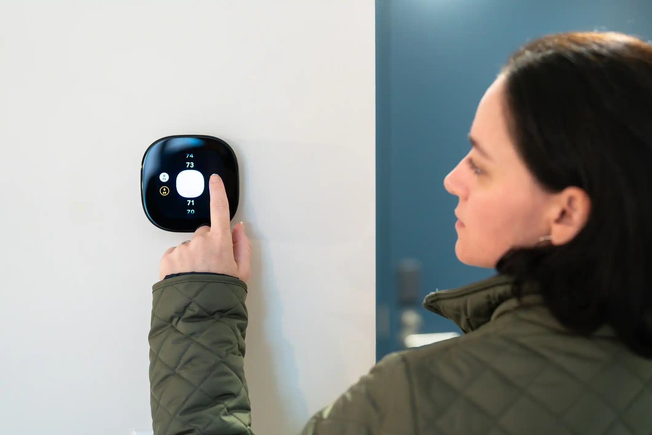 Woman pointing at a circular, black smart thermostat mounted on a white wall.