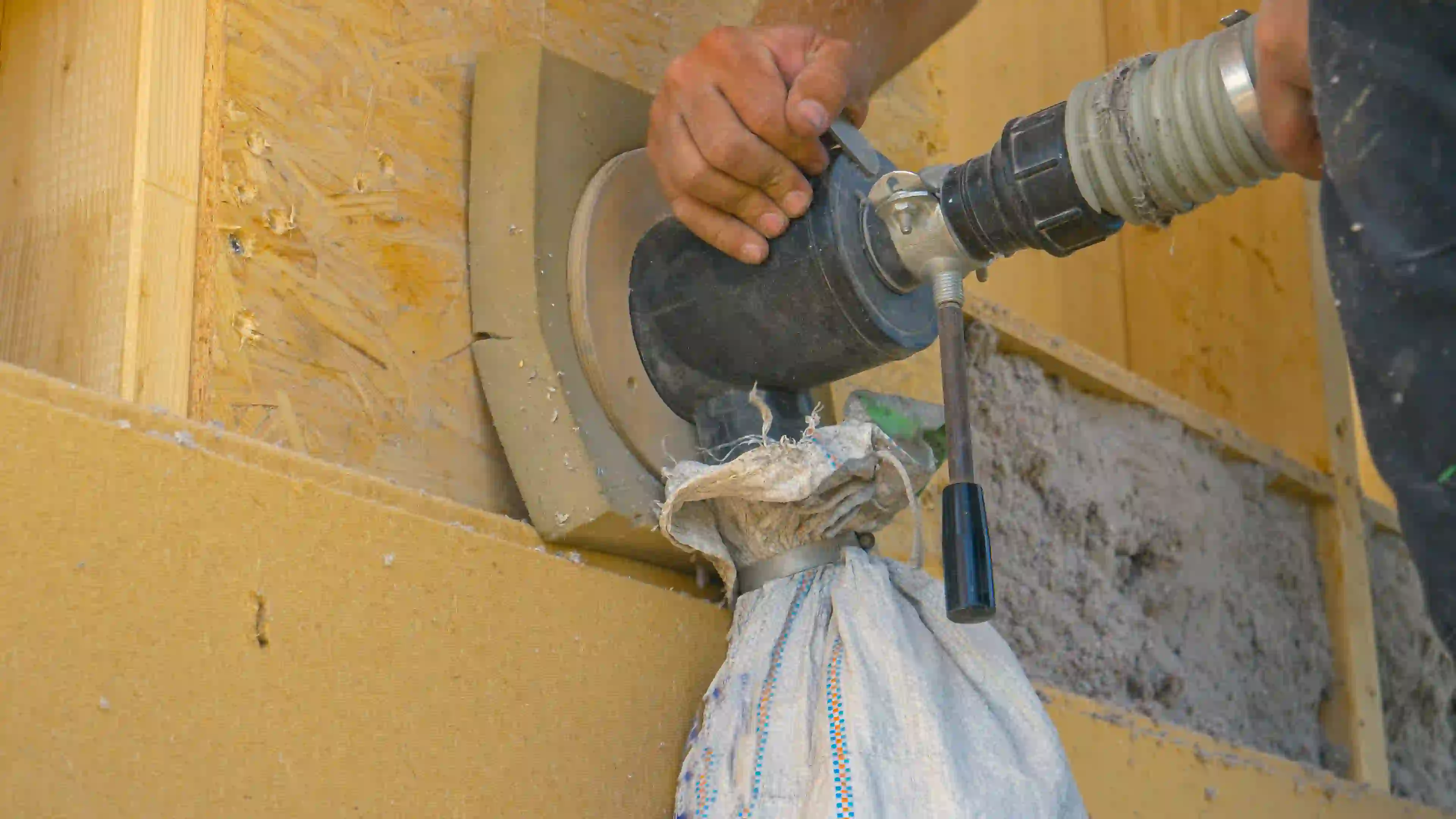 A close-up image shows a worker's hands operating a specialized machine for installing dense-pack insulation into a wall cavity. The machine features a nozzle and hose attachment and is sealed against the exterior of a wall, which is sheathed in OSB (Oriented Strand Board). A white cloth bag or screen, secured below the nozzle, is collecting excess grey loose-fill insulation (likely cellulose or fiberglass) that is being blown under high pressure into the sealed space. This technique is often used for insulating existing wall cavities.