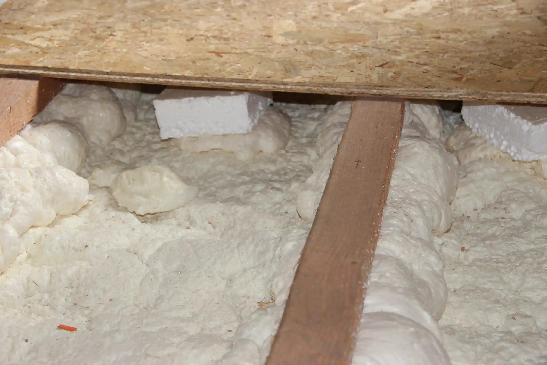 A close-up view shows a thick, dense application of spray foam insulation filling a floor cavity, likely in a crawl space. The off-white, expanded foam completely envelops the space around a central wooden floor joist. Two small, rectangular pieces of white expanded polystyrene (EPS) foam board are embedded in the foam near the top. Above the insulation, an OSB (Oriented Strand Board) subfloor or structural panel is visible, creating the ceiling of the insulated cavity. This image highlights the air-sealing and insulating qualities of spray foam.