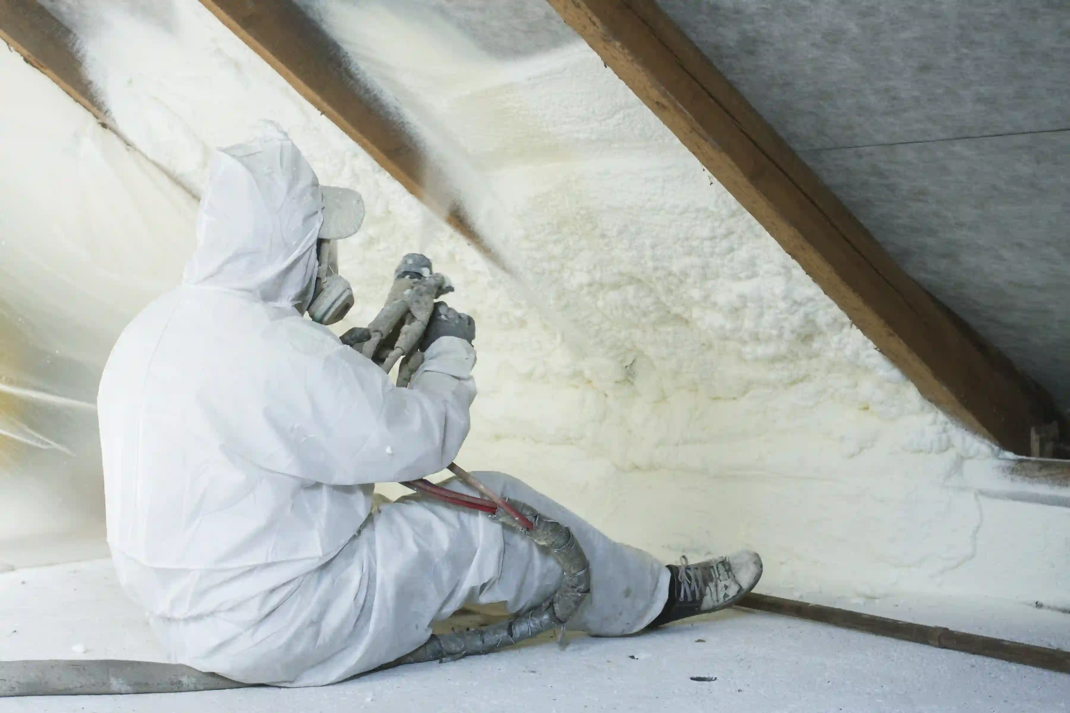 A person wearing a full white protective suit, hood, and respirator mask is applying spray foam insulation to the underside of a sloped roof or rafter wall.